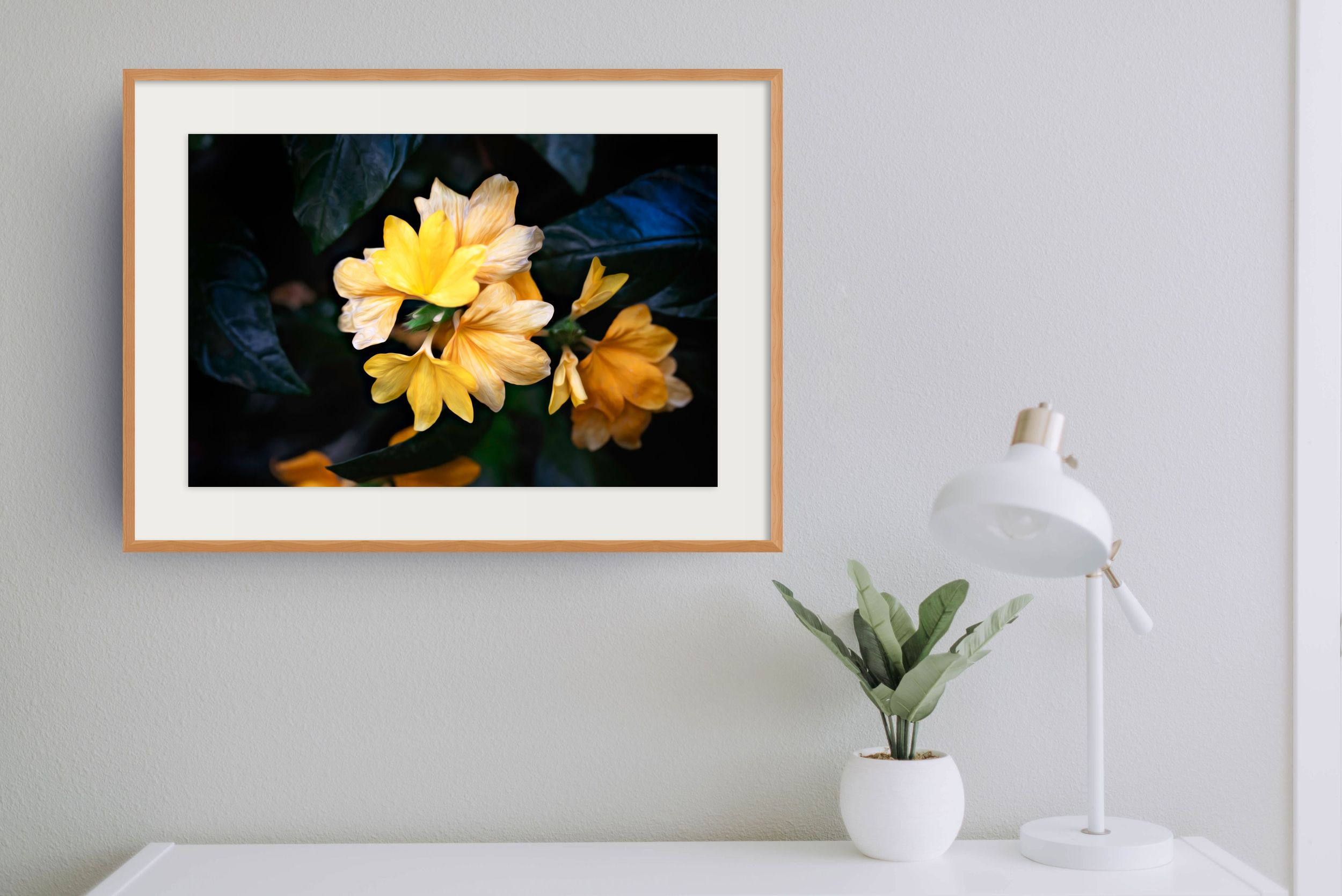 Framed yellow flower photography print with dark background, displayed above a white desk with a small plant and white lamp.