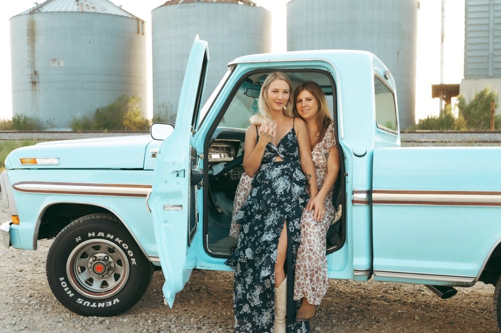 Two women in floral maxi dresses pose beside a vintage light blue pickup truck near grain silos.
