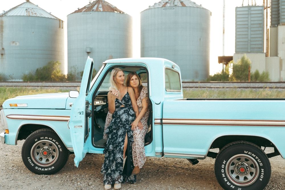 Two women in floral maxi dresses pose by a vintage light blue pickup truck with grain silos in the background.