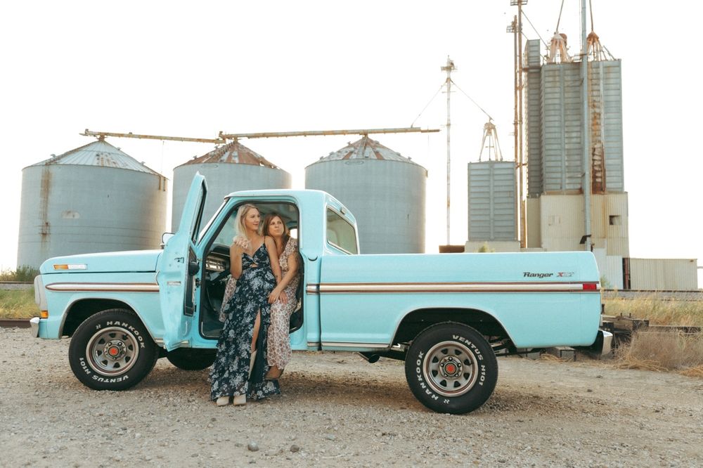 Two women in floral dresses pose beside a classic light blue vintage pickup truck near grain silos.