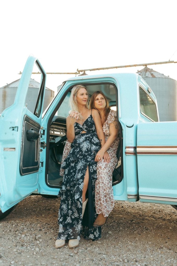 Two women in floral maxi dresses pose beside a vintage turquoise pickup truck on a gravel lot.