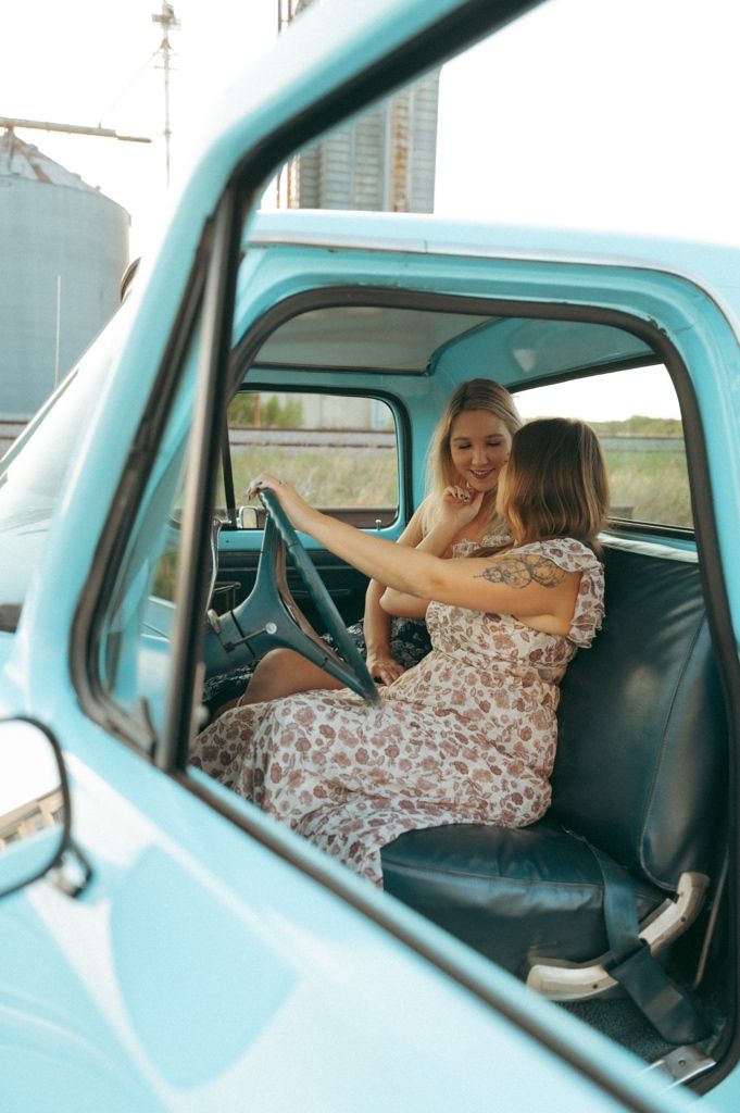 Two women in floral dresses sitting inside a vintage turquoise pickup truck, smiling and interacting in Prosper TX