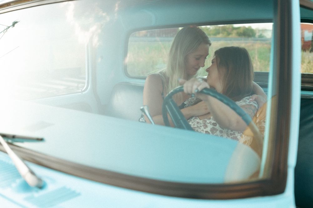 Two women sharing an intimate moment inside a vintage car, viewed through the windshield on a sunny day in Prosper TX