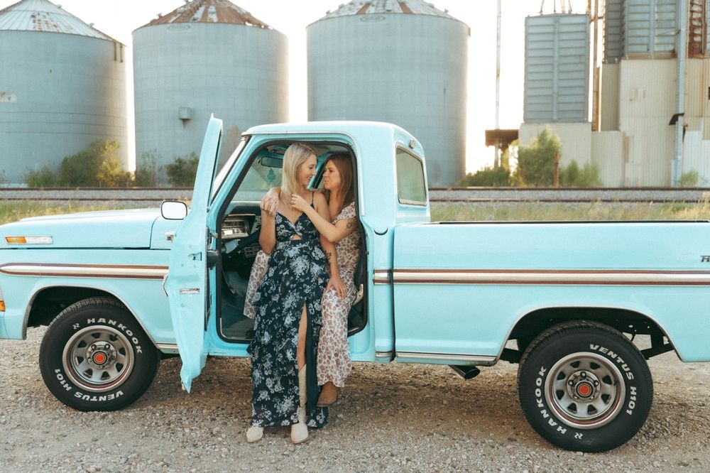 Two women in floral outfits pose beside a vintage light blue pickup truck near grain silos at golden hour in Prosper TX