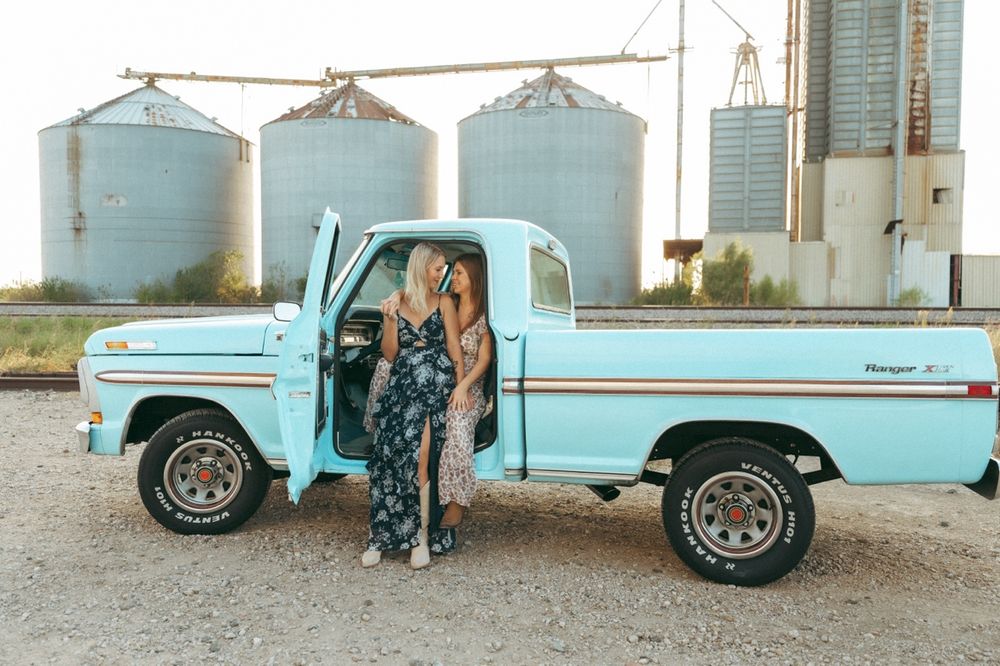 Two women in floral outfits pose beside a vintage light blue Ford pickup truck near grain silos at golden hour in Prosper TX