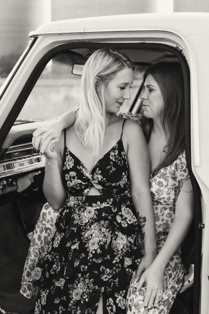 Two women in floral dresses pose together by a vintage white truck in a black and white photo.