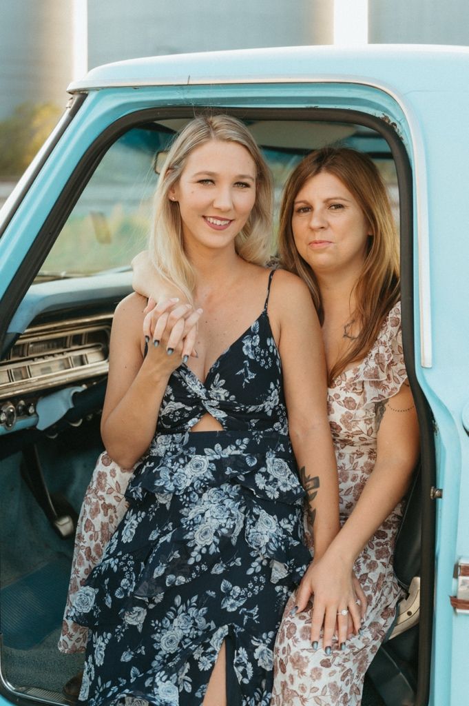 Two young women in floral dresses pose beside a vintage light blue pickup truck outdoors.