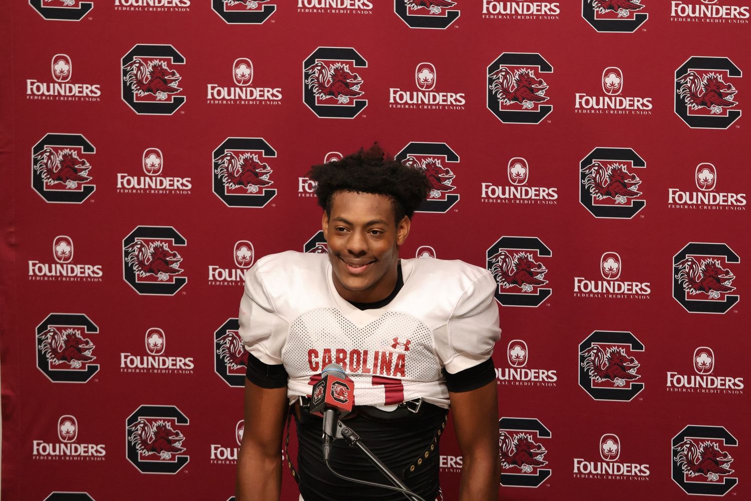 South Carolina football player in uniform smiles at a press conference podium.