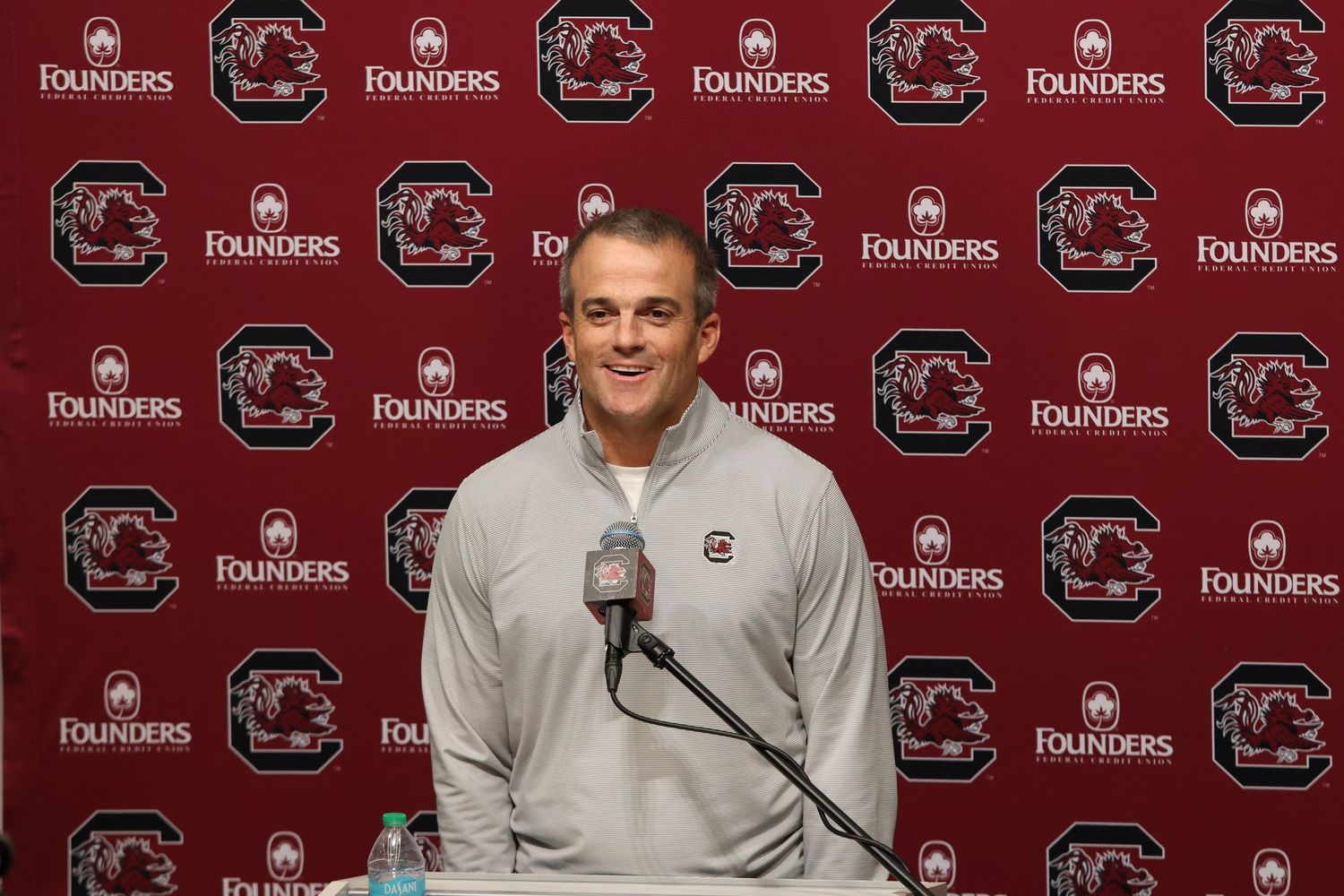 South Carolina football coach smiles while speaking at a press conference podium.