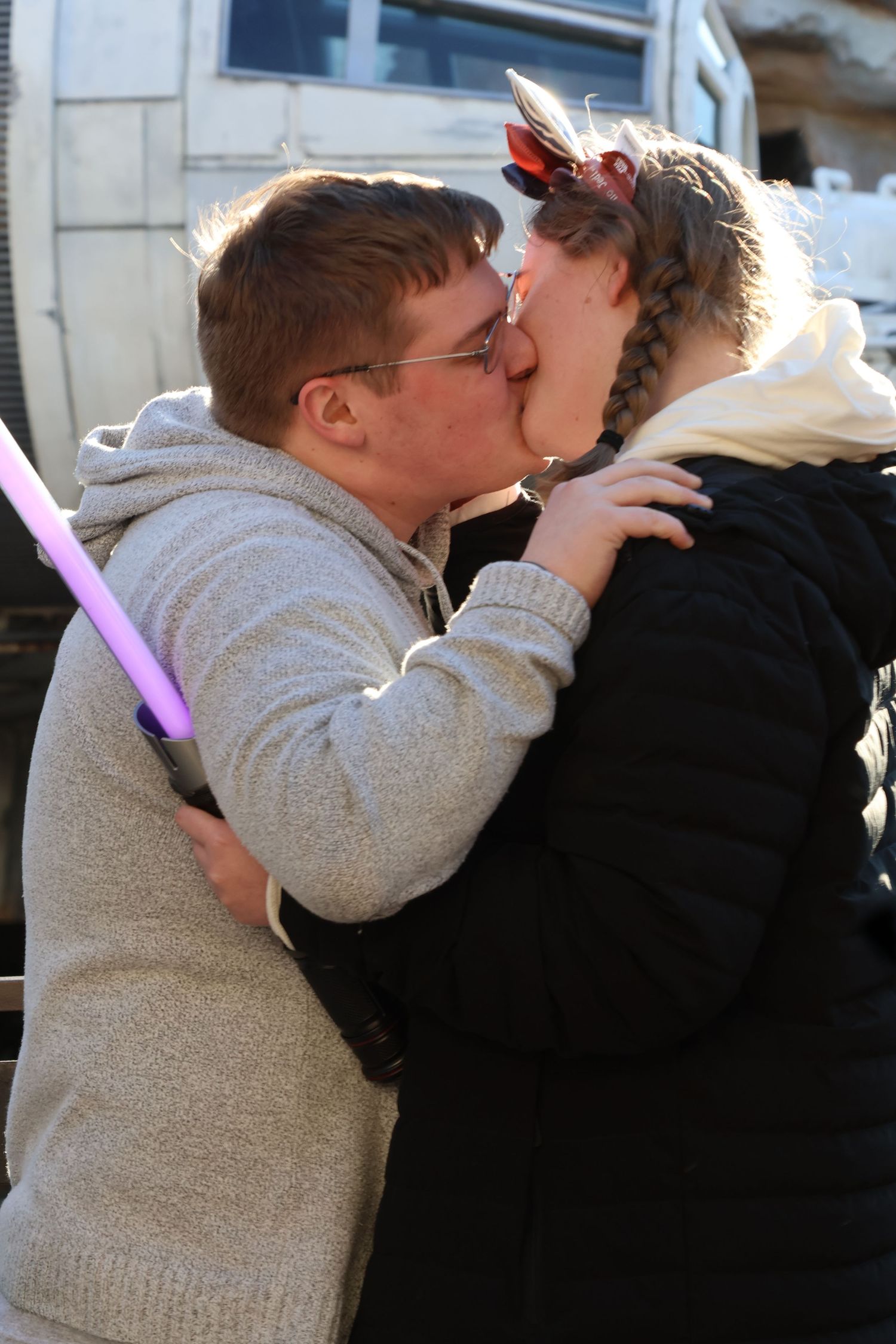 Couple kisses outdoors while holding a glowing pink toy lightsaber.