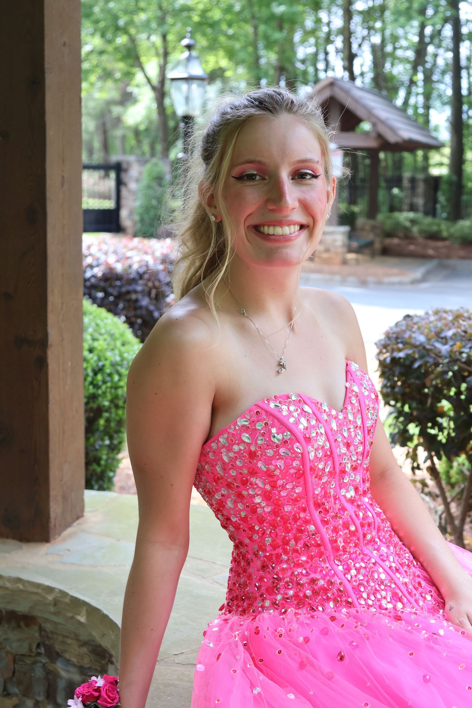 Teen in a jeweled bright pink prom gown smiles beside a wooden pavilion.
