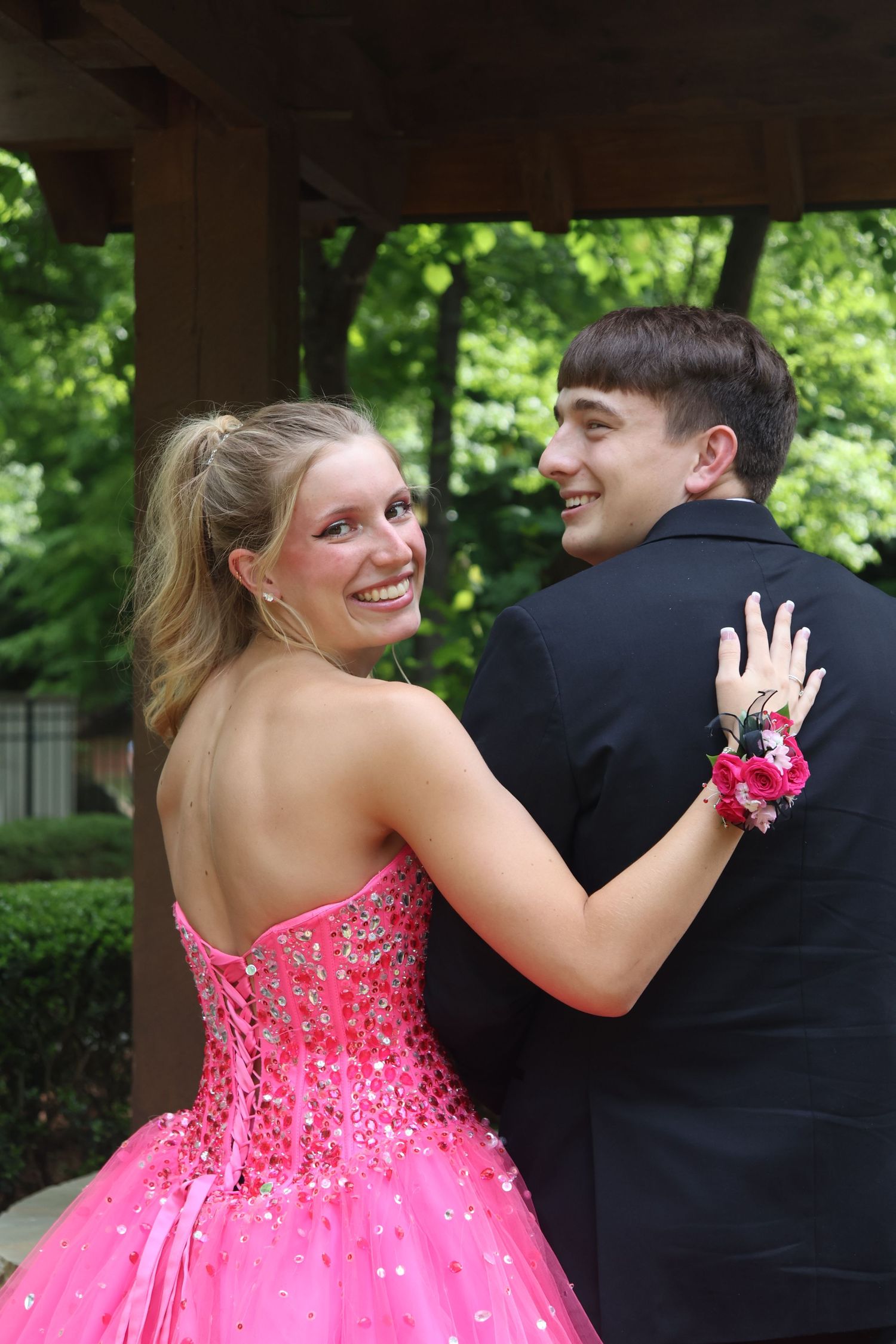 Prom couple looks back at the camera while walking beneath a wooden pavilion.