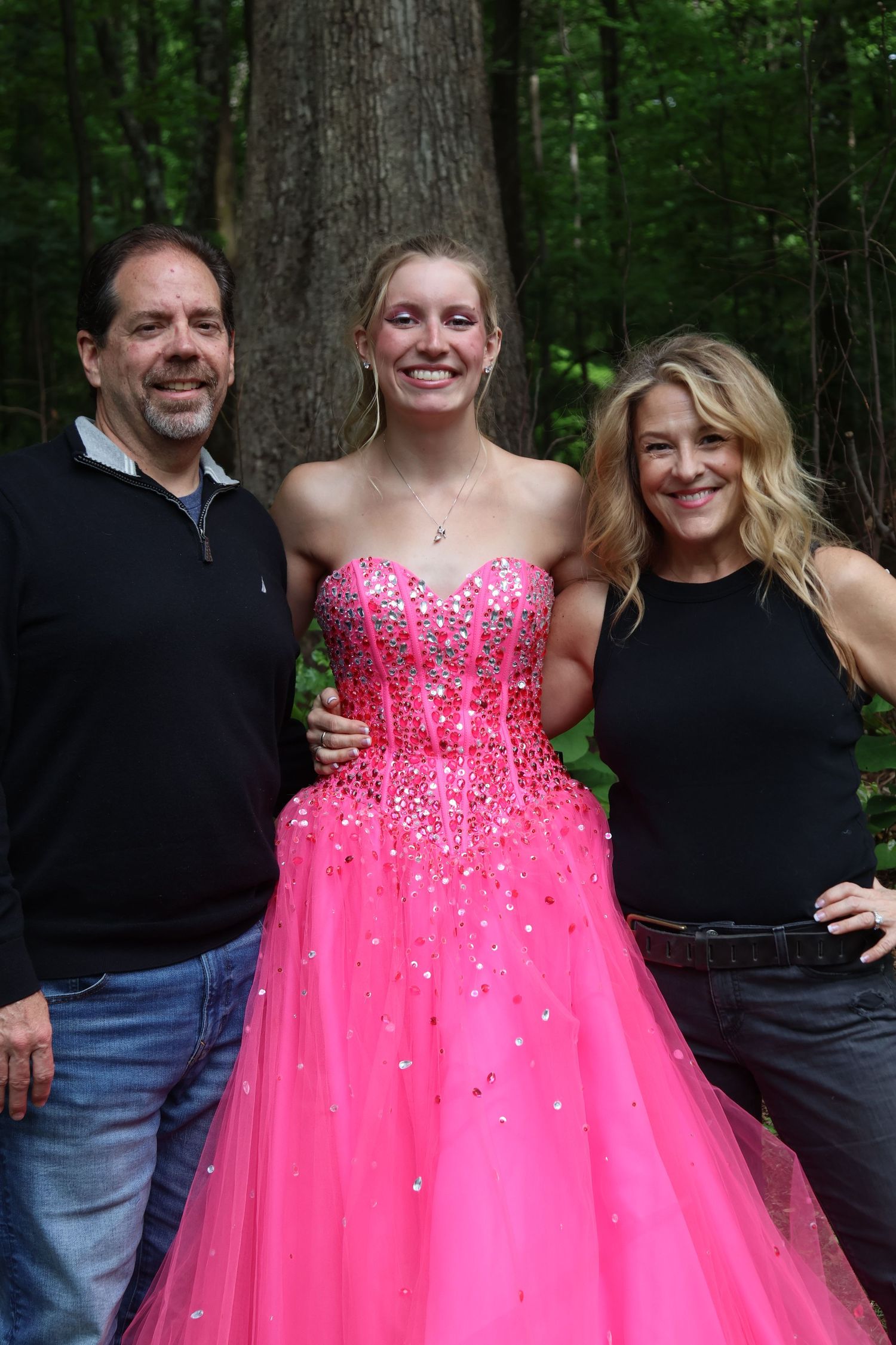 Teen in a bright pink prom gown poses outdoors with her parents.