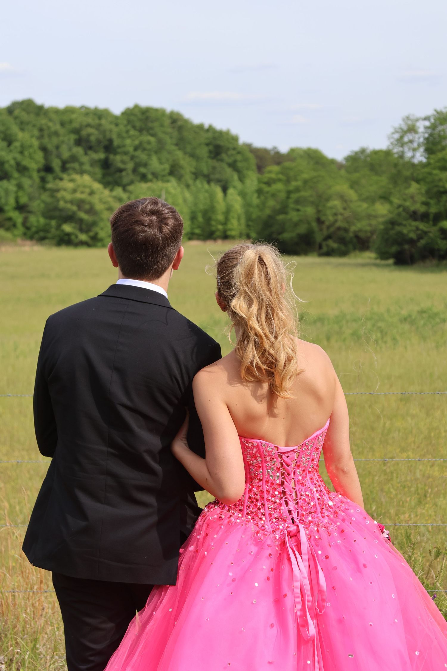 Prom couple in a black suit and bright pink gown stand together facing a grassy field.
