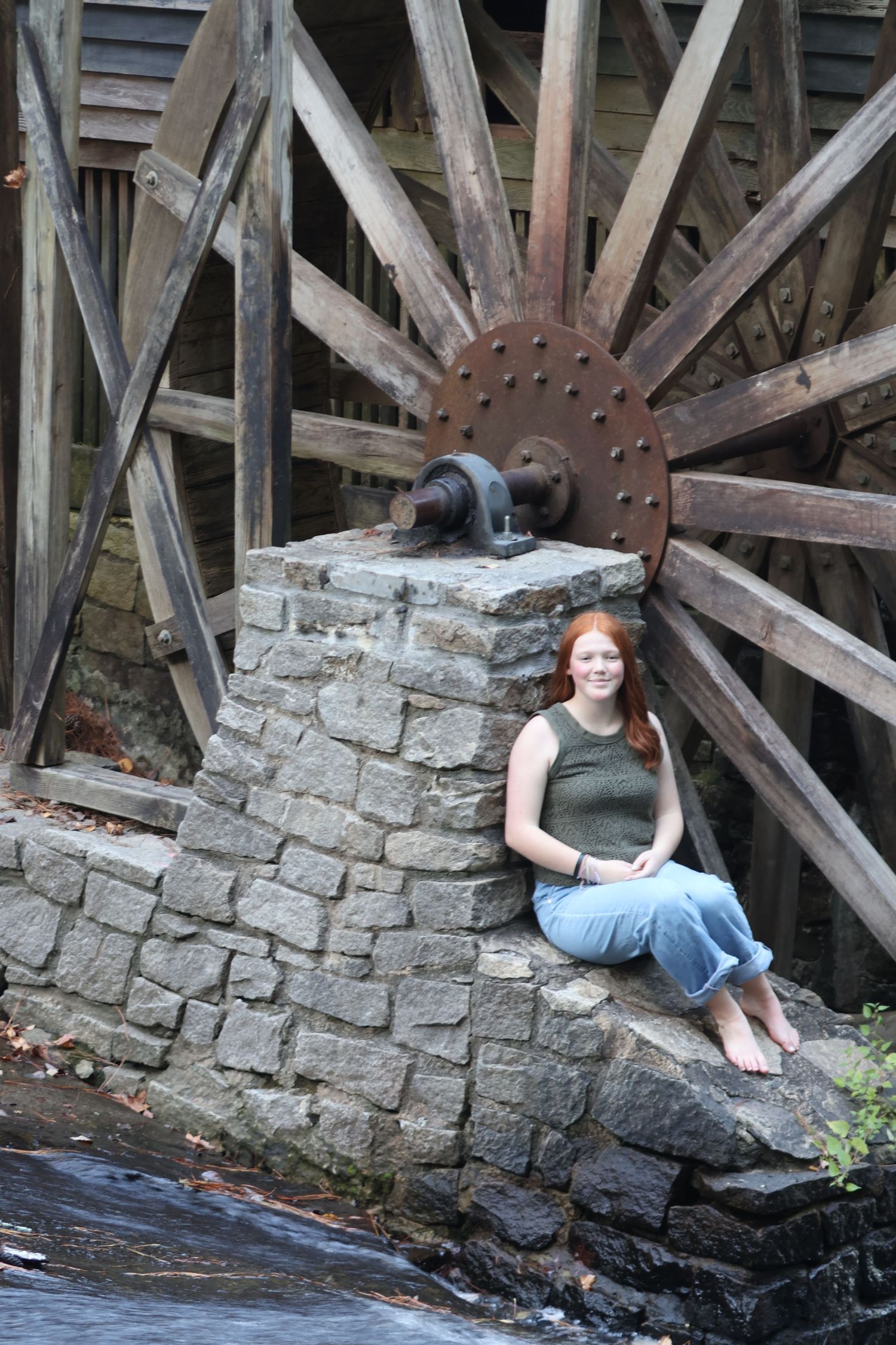 Red-haired young woman sits on a stone ledge beside a large wooden waterwheel.