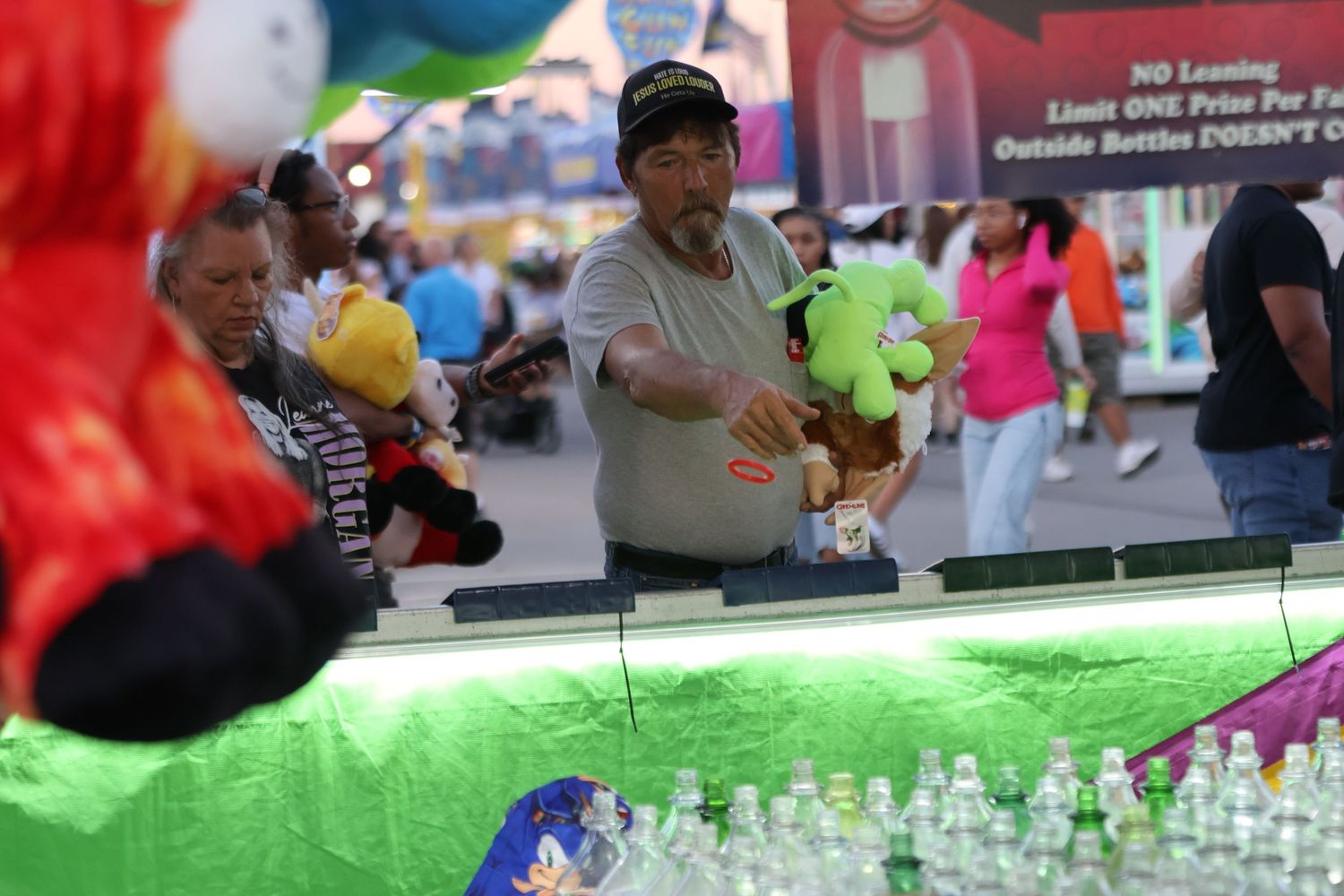 Carnival game attendant stands behind a counter lined with bottles and colorful prizes.