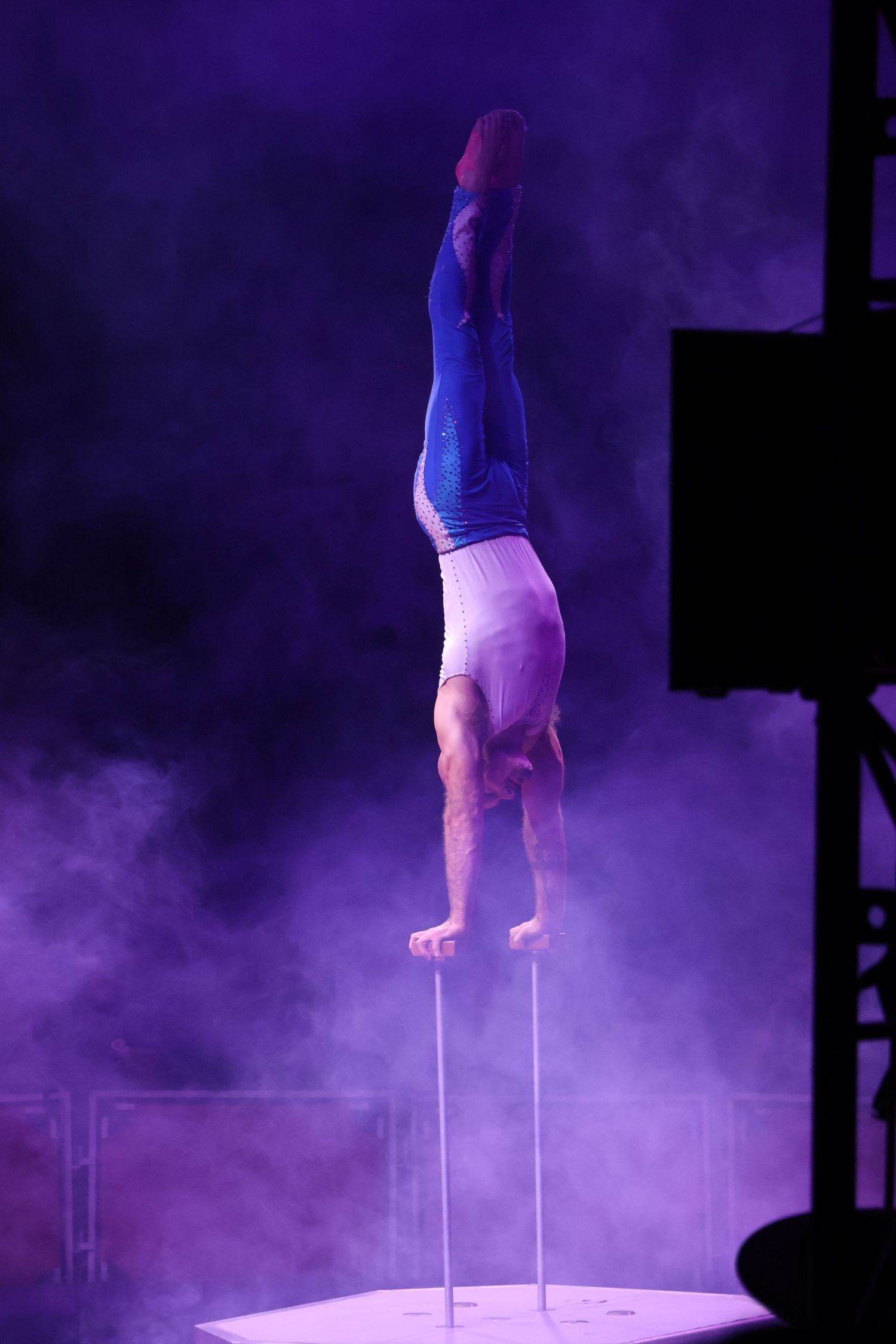 Acrobat balances in a handstand on canes amid purple stage light and fog.