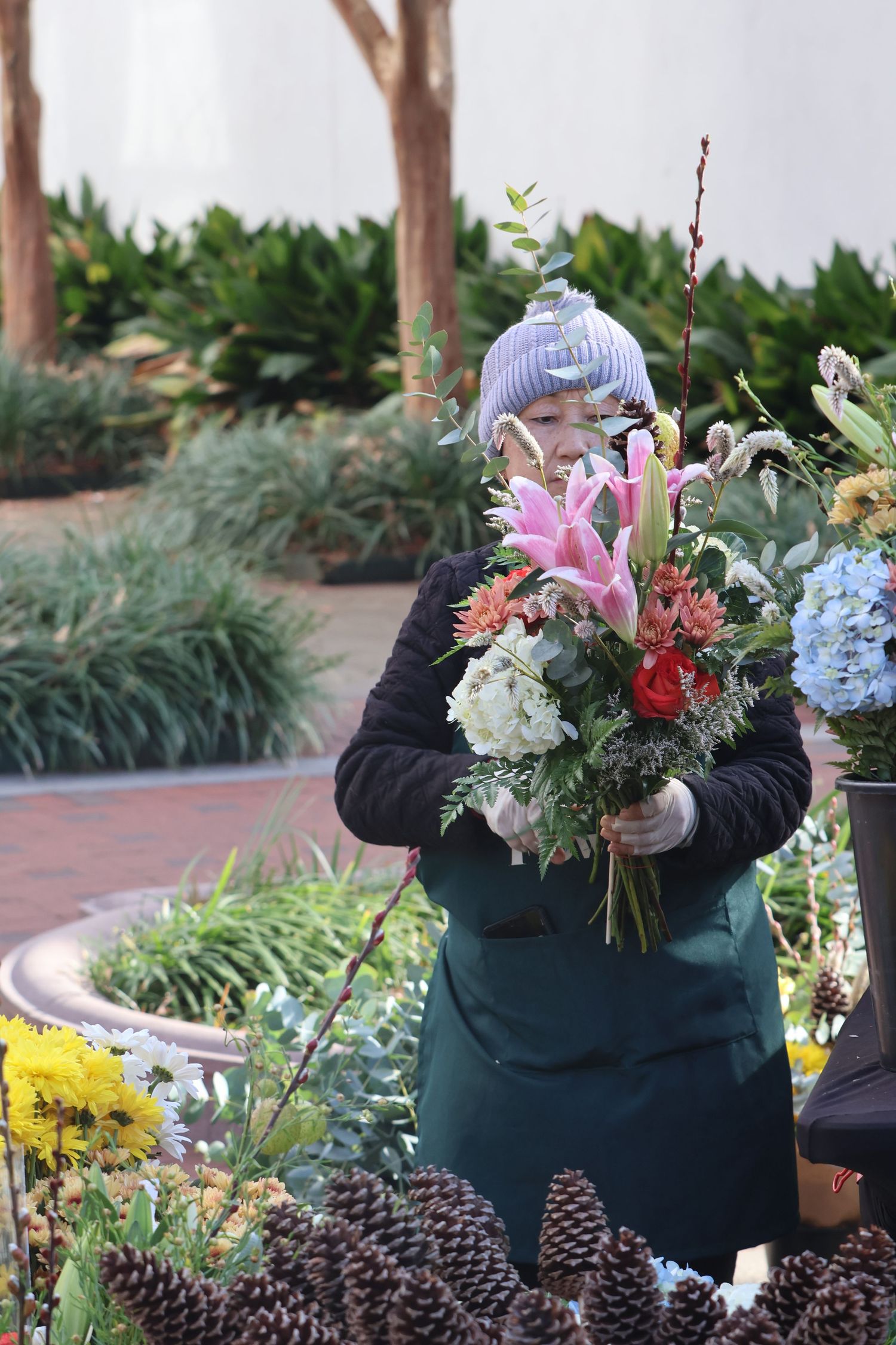 Flower vendor wearing a knit hat arranges a large mixed bouquet at an outdoor market.
