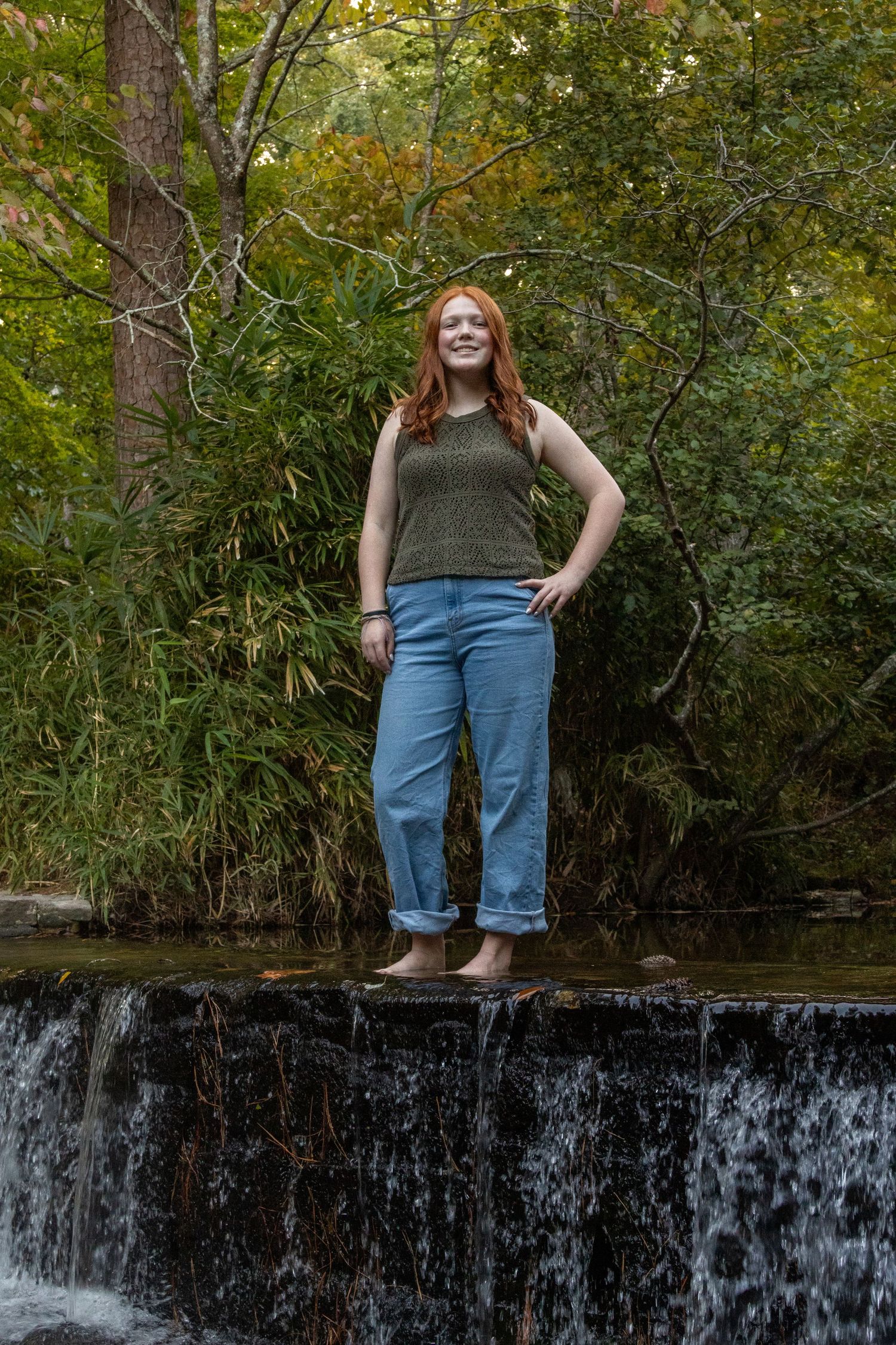 Red-haired young woman stands barefoot on the edge of a small waterfall in the woods.