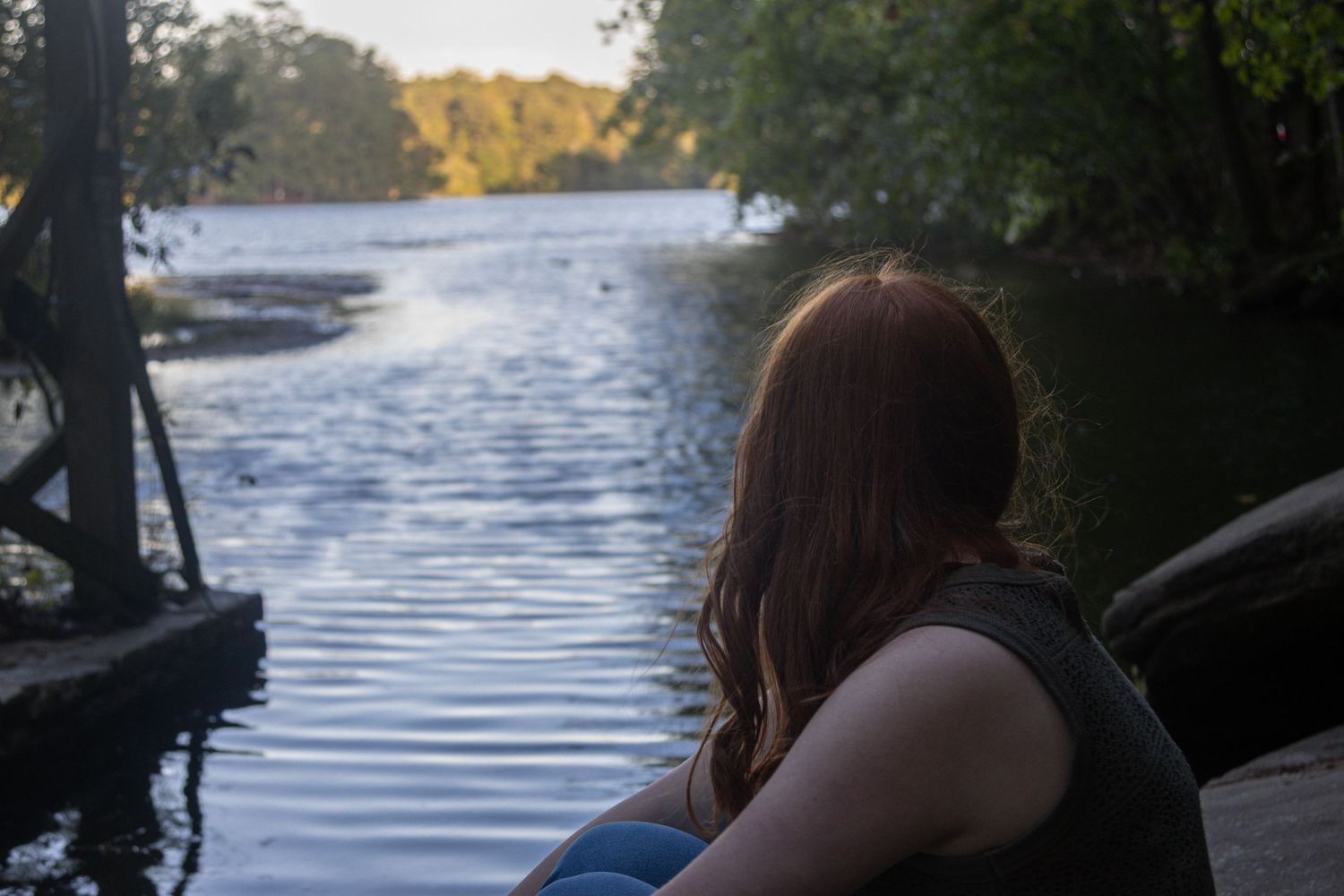 Young woman sits in silhouette looking across a calm river at sunset.