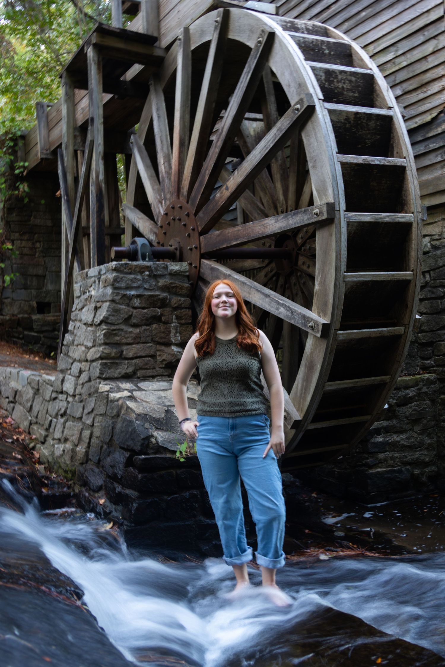 Young woman stands barefoot in flowing water in front of a large wooden waterwheel.
