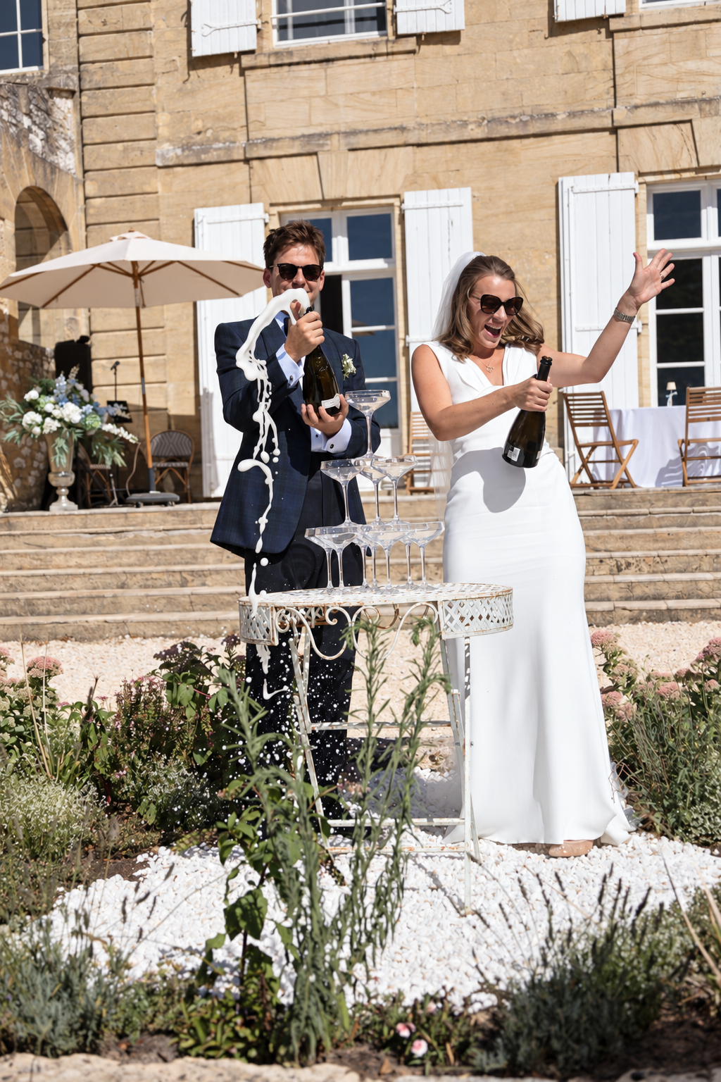 Beth and Matt opening champagne bottles beside a tower of glasses in the courtyard at Château Sainte-Croix.