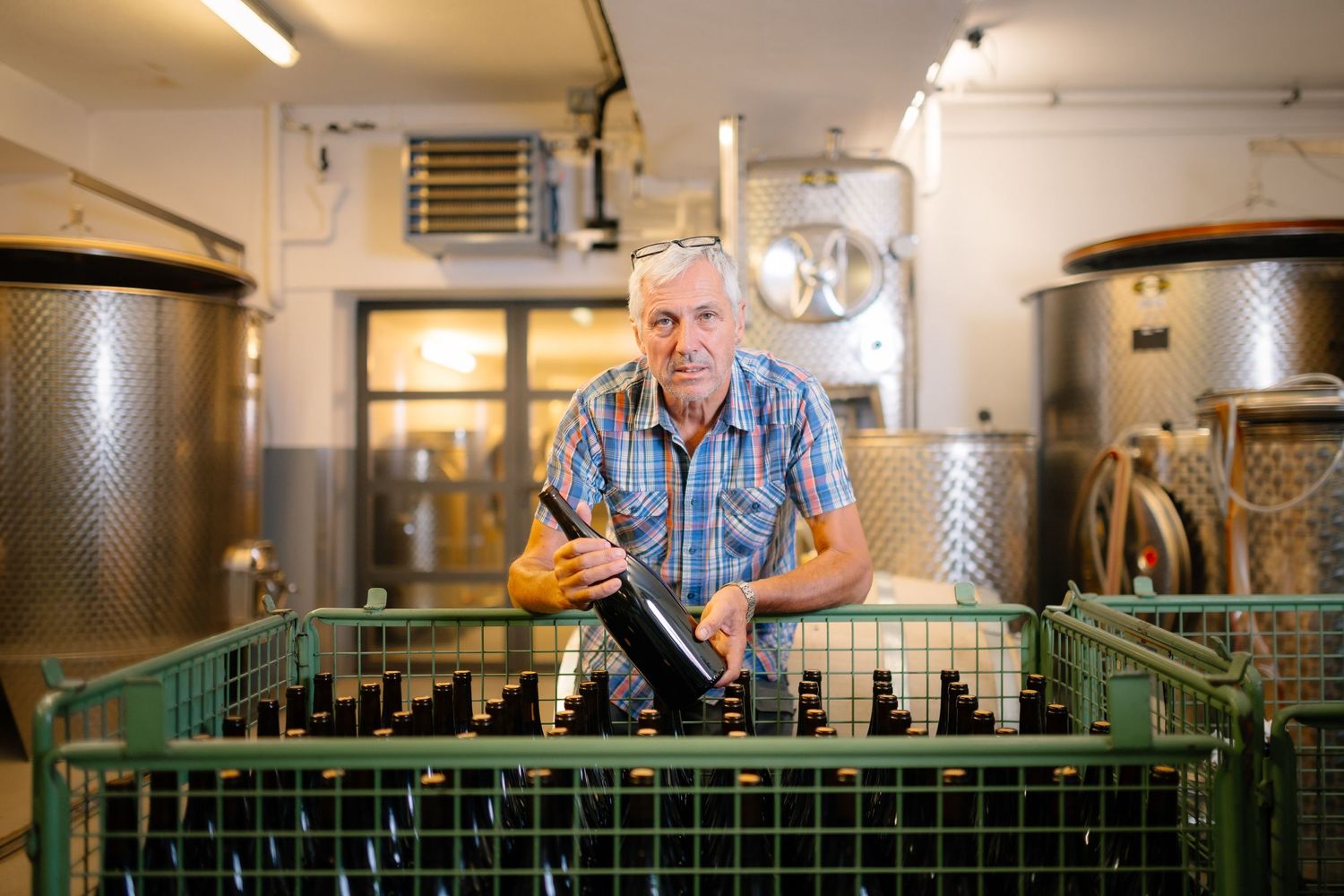 Worker handling wine bottles in industrial bottling facility with stainless steel tanks.