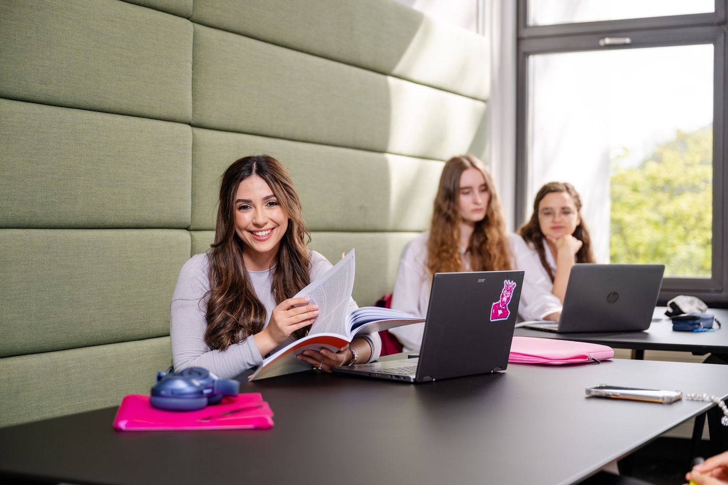 Students studying together at modern workspace with laptops and pink accessories.