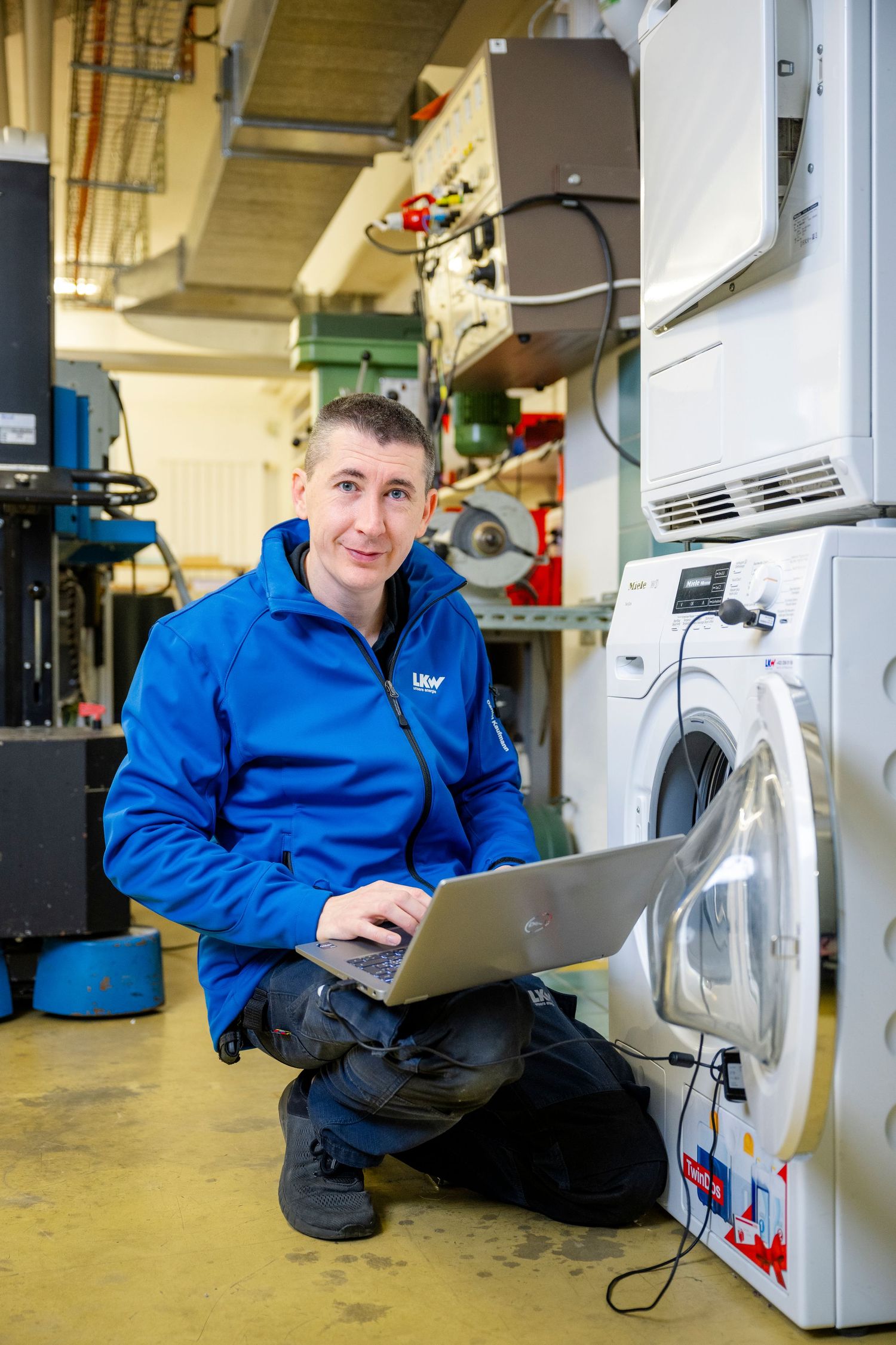 Maintenance worker in blue jacket using laptop while servicing appliances.
