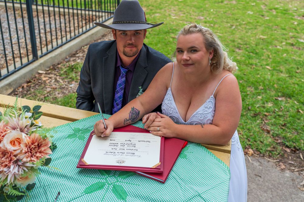 Couple signing marriage documents at outdoor ceremony with floral decorations.