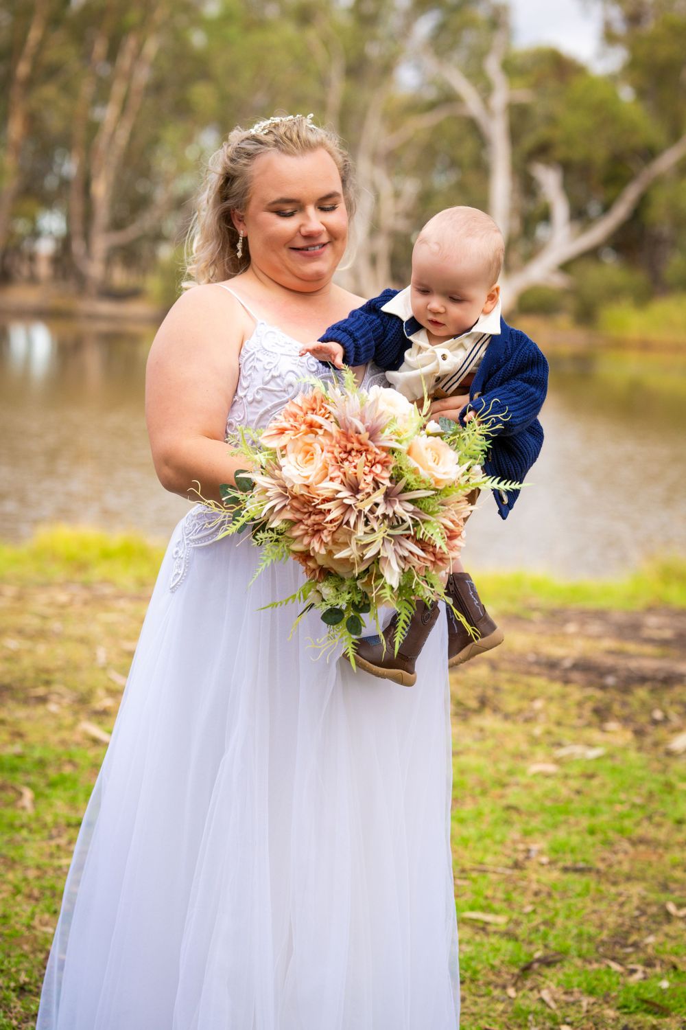 A tender moment with a baby in formal wear and beautiful bouquet by a lake.