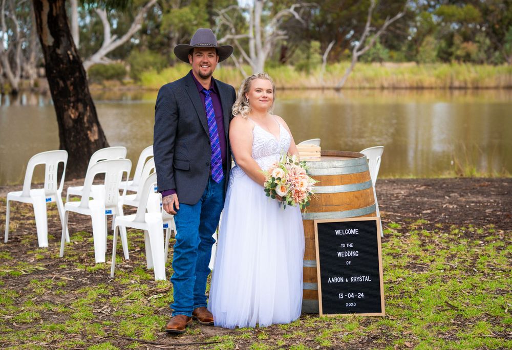 A rural wedding ceremony setup with white chairs and chalkboard sign near a serene lake.
