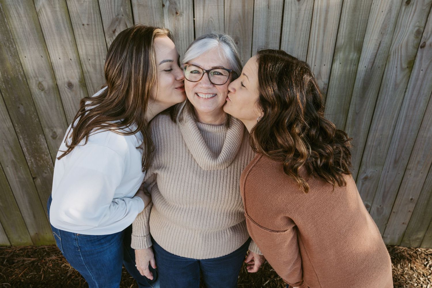 Three generations share a tender family moment against wooden fence backdrop.