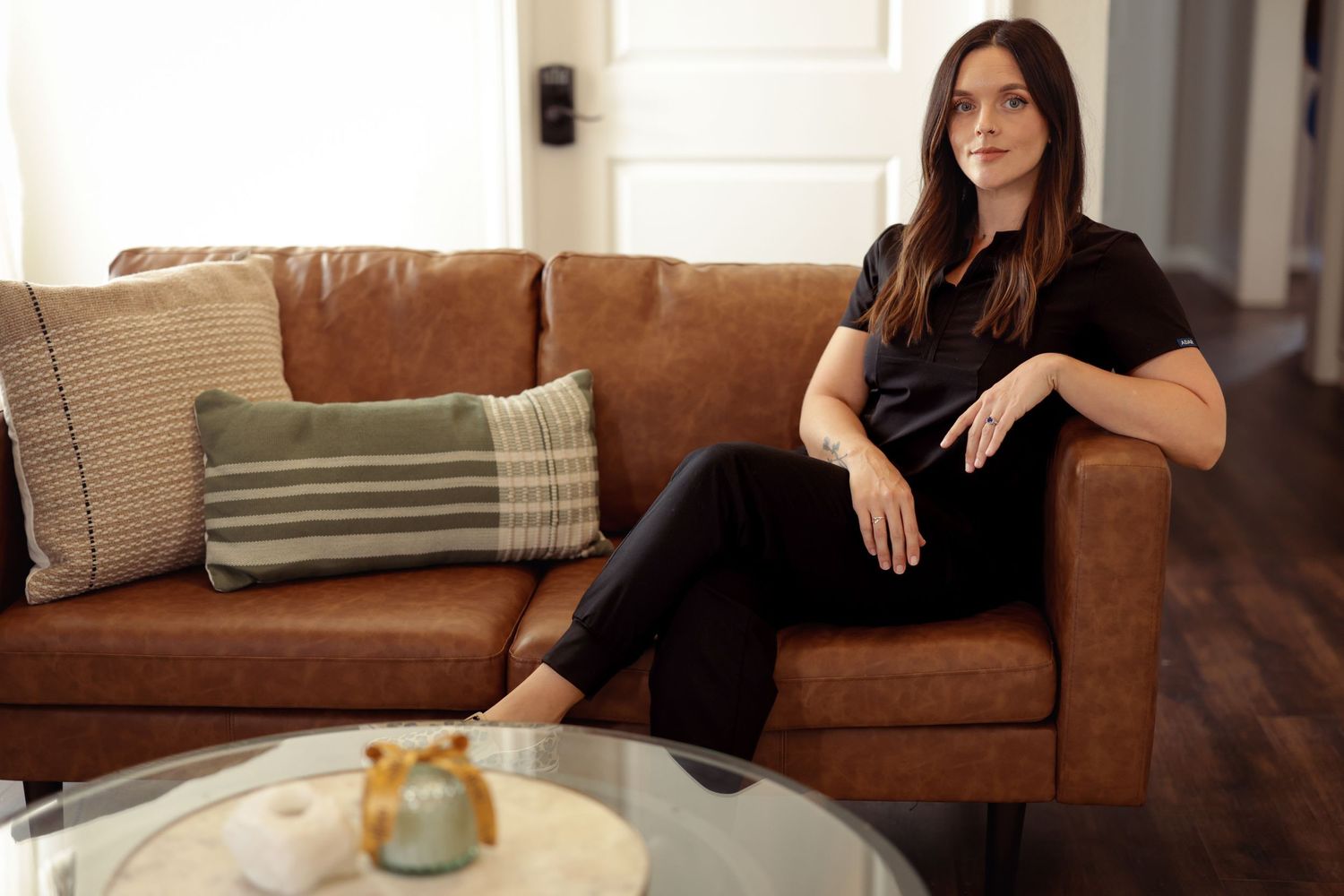 A woman in black lounges comfortably on a tan leather couch next to a coffee table in a modern living room. An esthetician's branding.