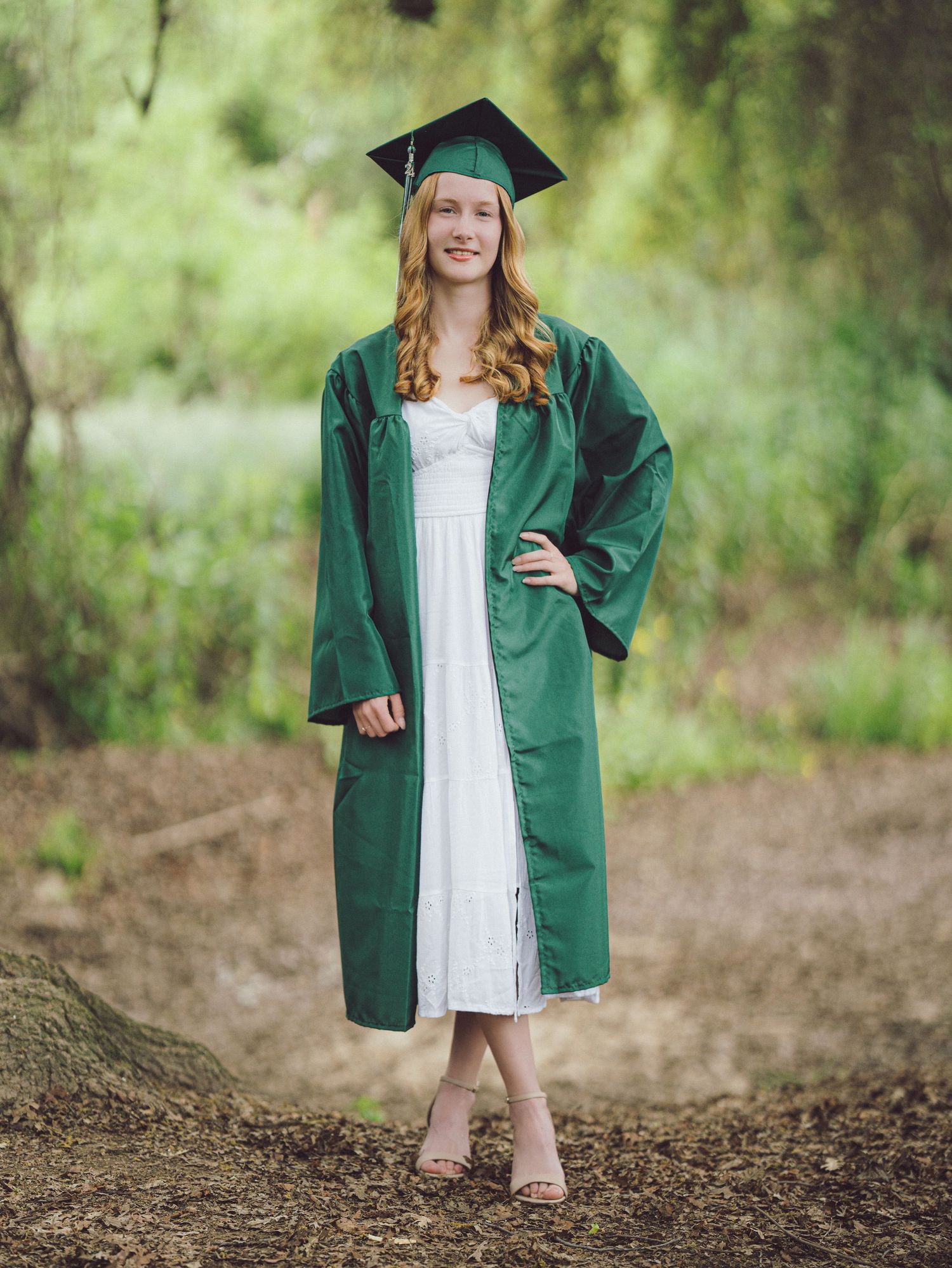 Smiling graduate wearing a green cap and gown over a white dress standing outdoors in nature.