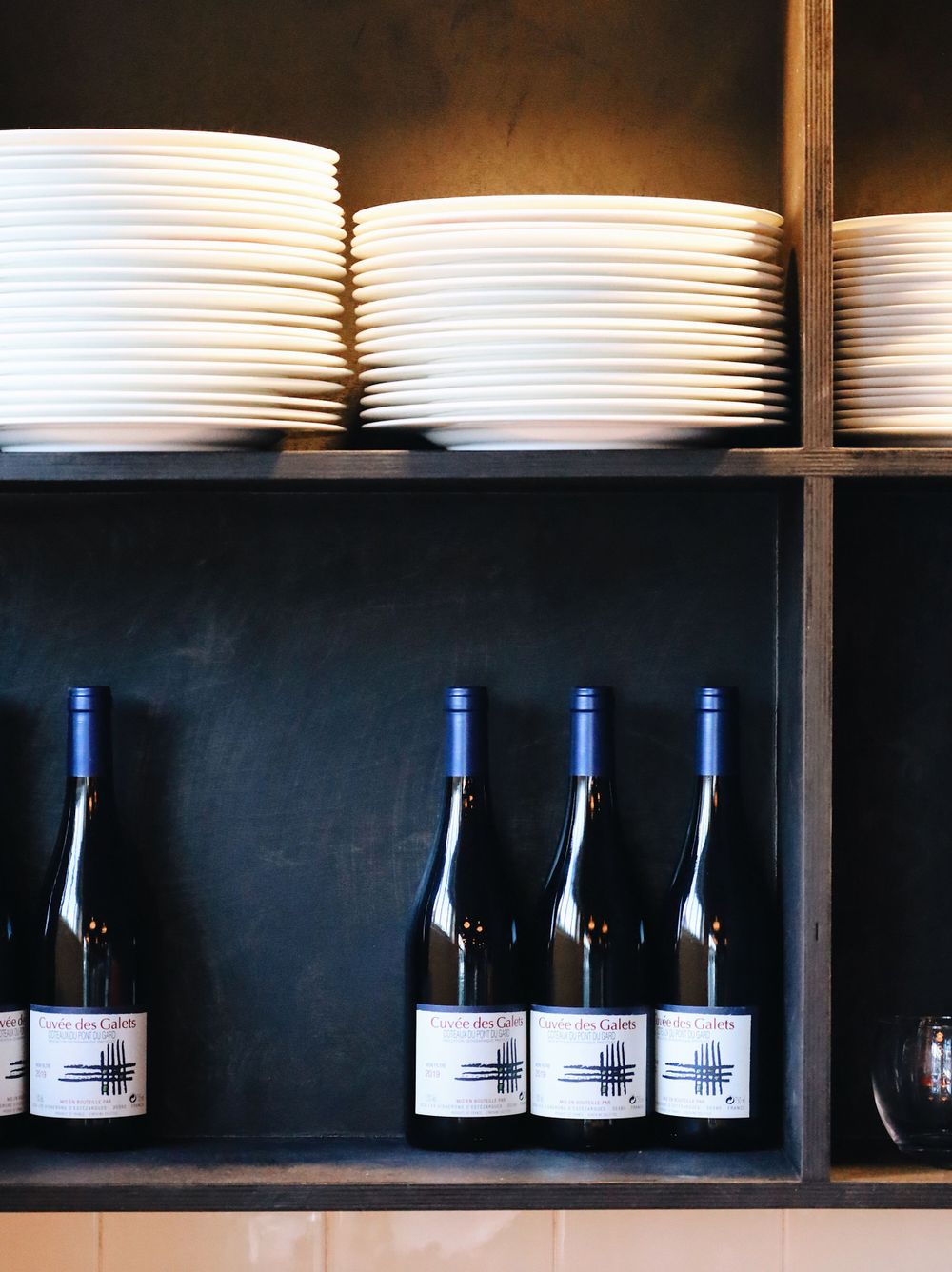Wine bottles displayed on dark shelves with white plates stacked above.