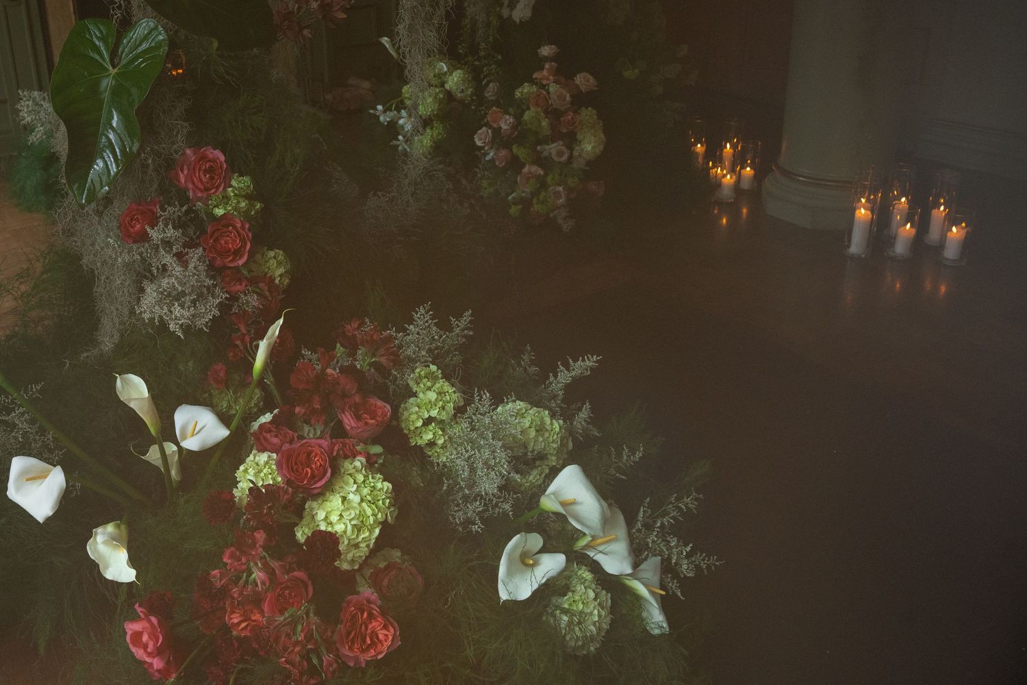 Red flowers and white calla lilies arranged with greenery against a dark background with glowing candles.