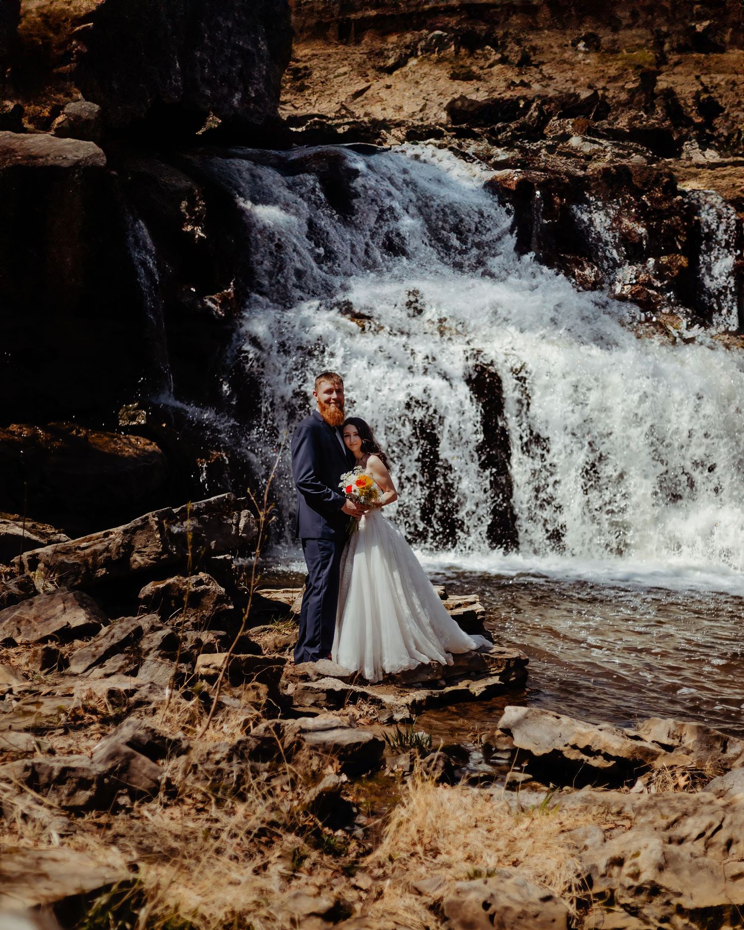 Wedding couple poses in front of rushing waterfall on rocky terrain.