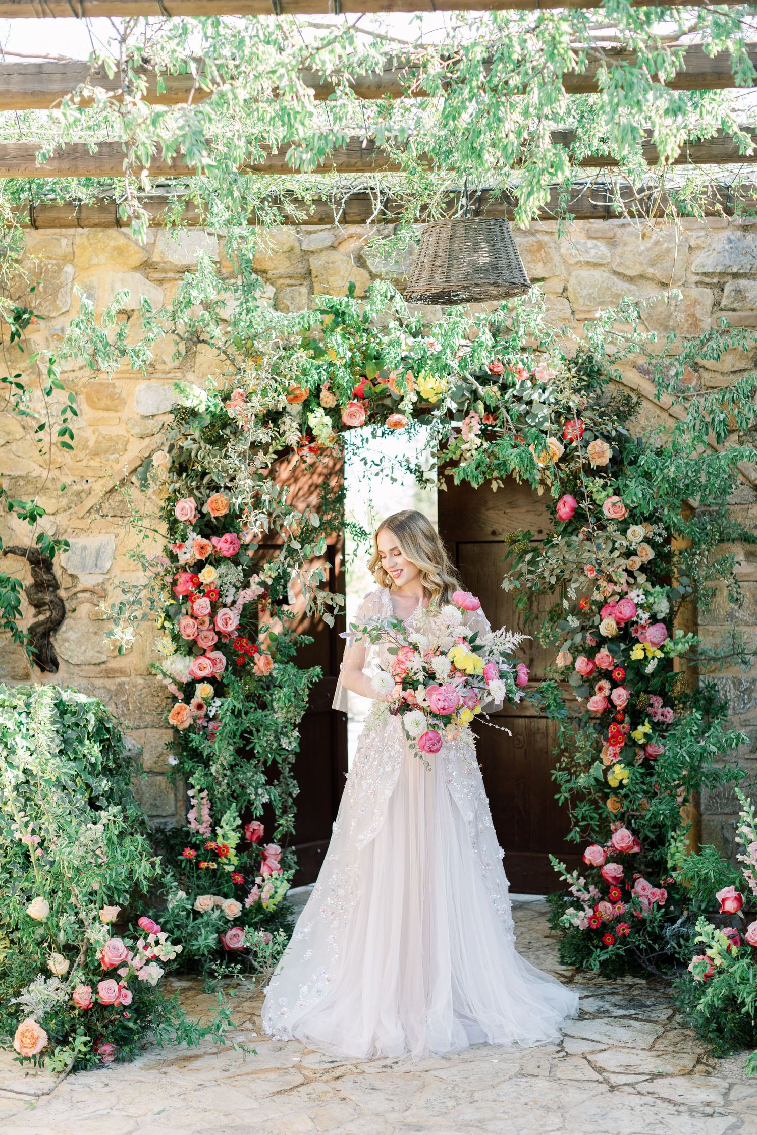 A bride in a flowing white gown stands beneath a romantic archway adorned with climbing roses and greenery.