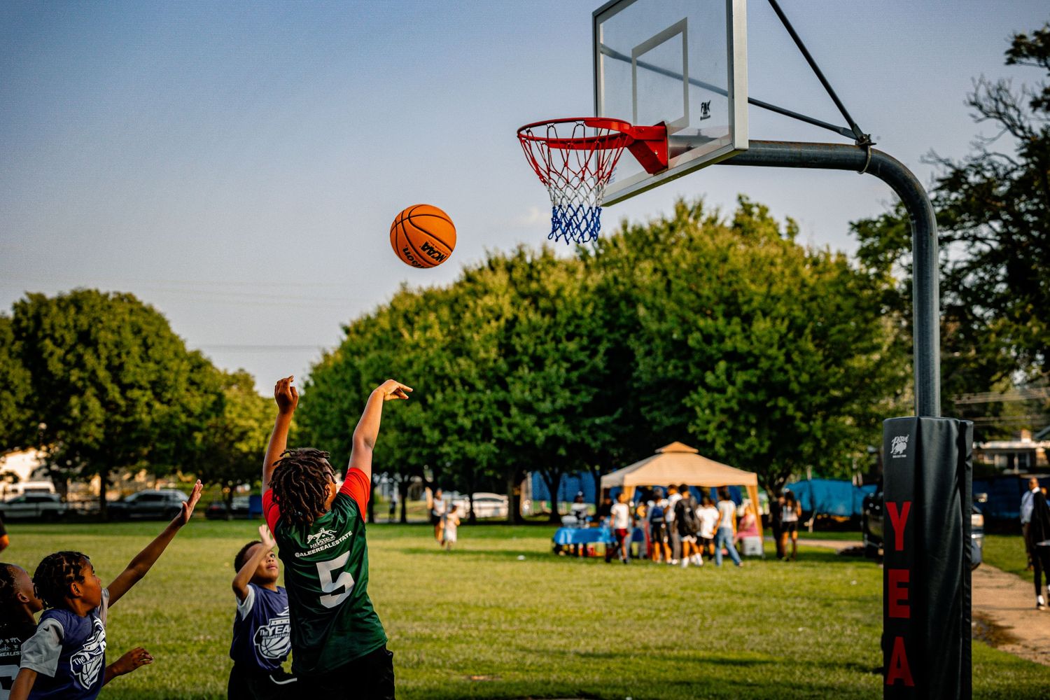 Orange basketball soars towards hoop during outdoor game with trees in background.