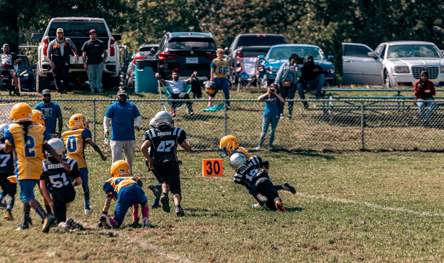 Youth football players compete on a field with parked cars and spectators in the background.