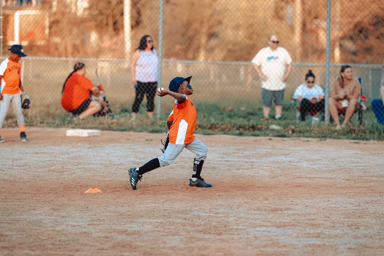 Youth baseball players in orange and blue uniforms practicing on a dirt field at sunset.
