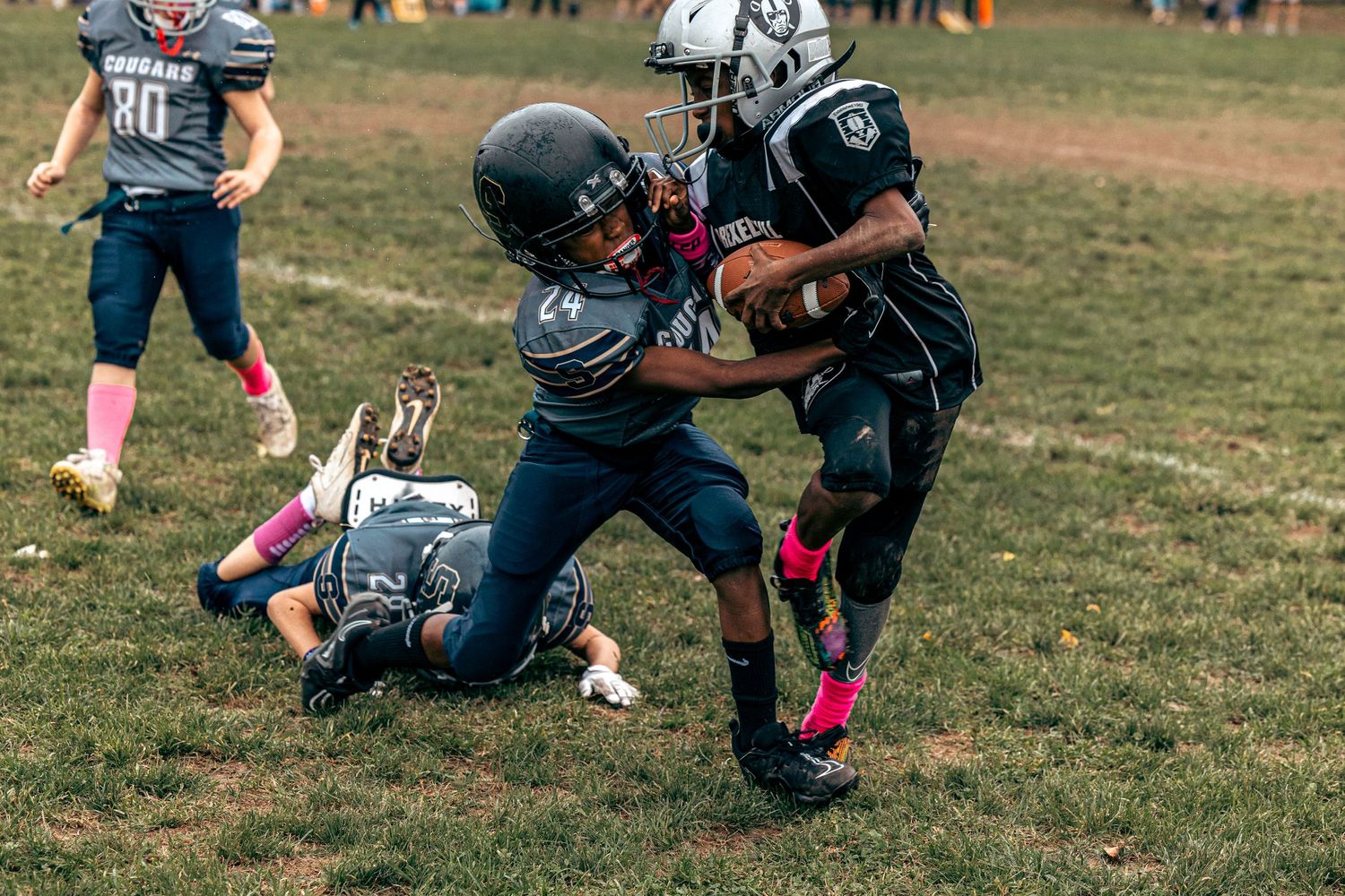 Youth football players in dark uniforms with pink accents engage in a tackle on the field.
