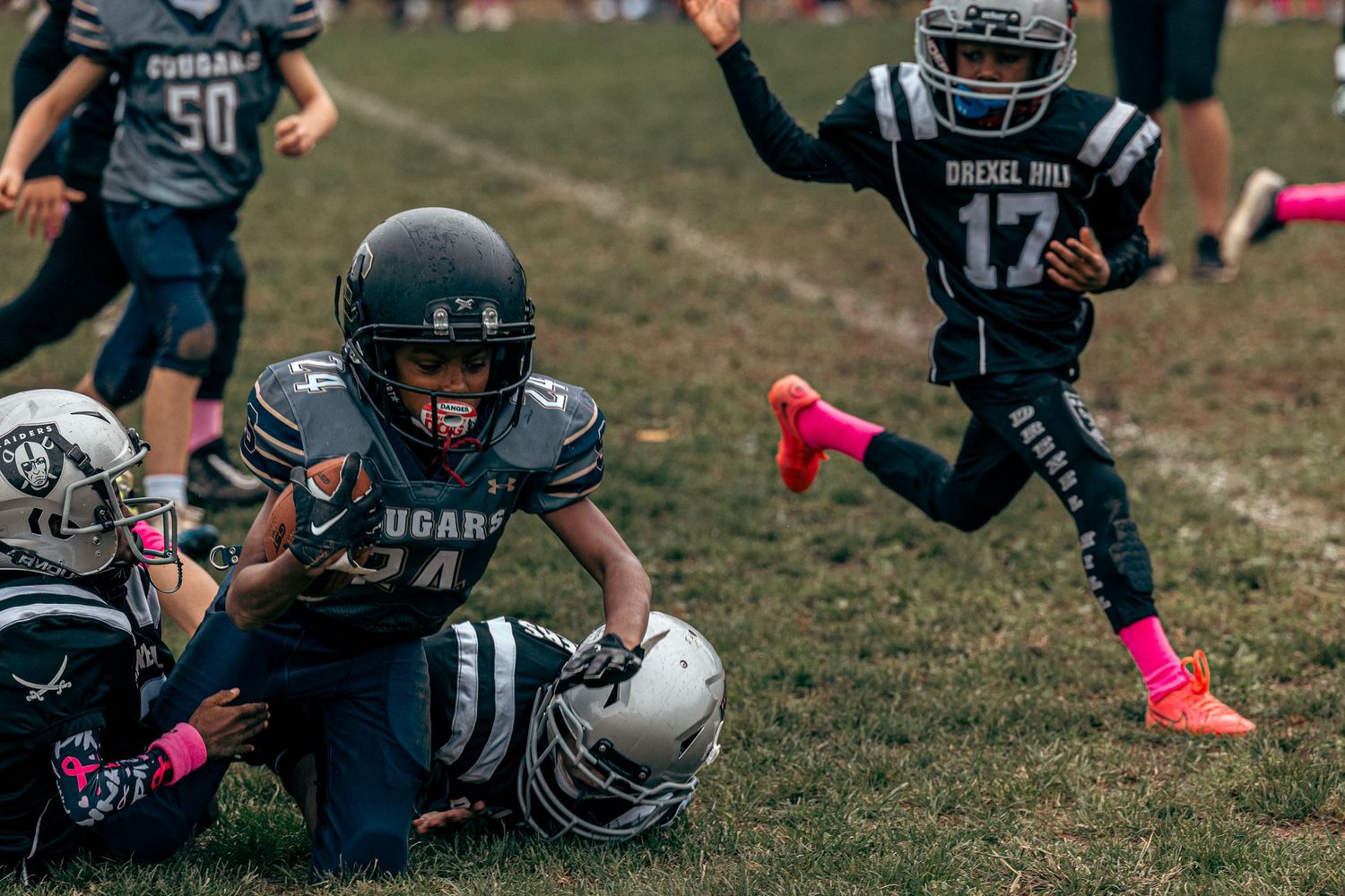 Youth football players in dark uniforms with pink cleats tackle during an intense game play on a grass field.