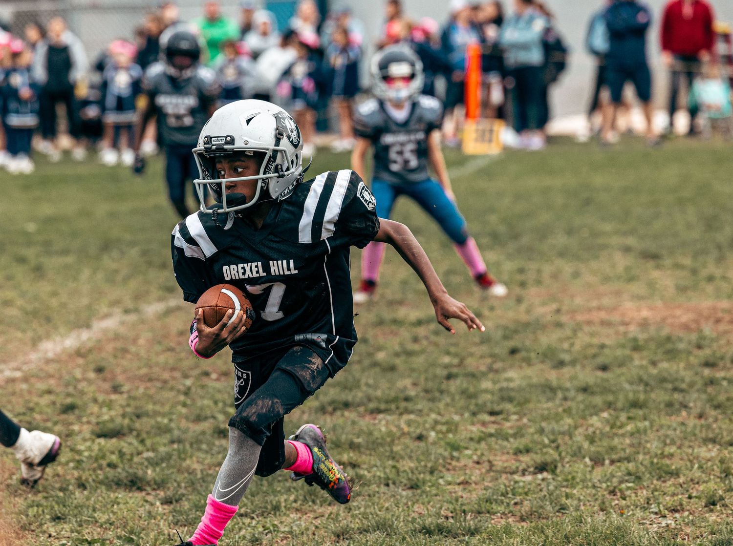 Young football player in dark uniform with pink cleats runs with the ball during a game.