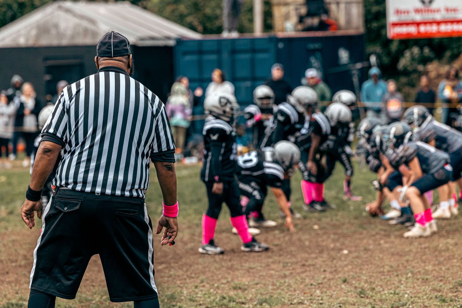 Referee oversees youth football players lined up in formation wearing pink accessories.
