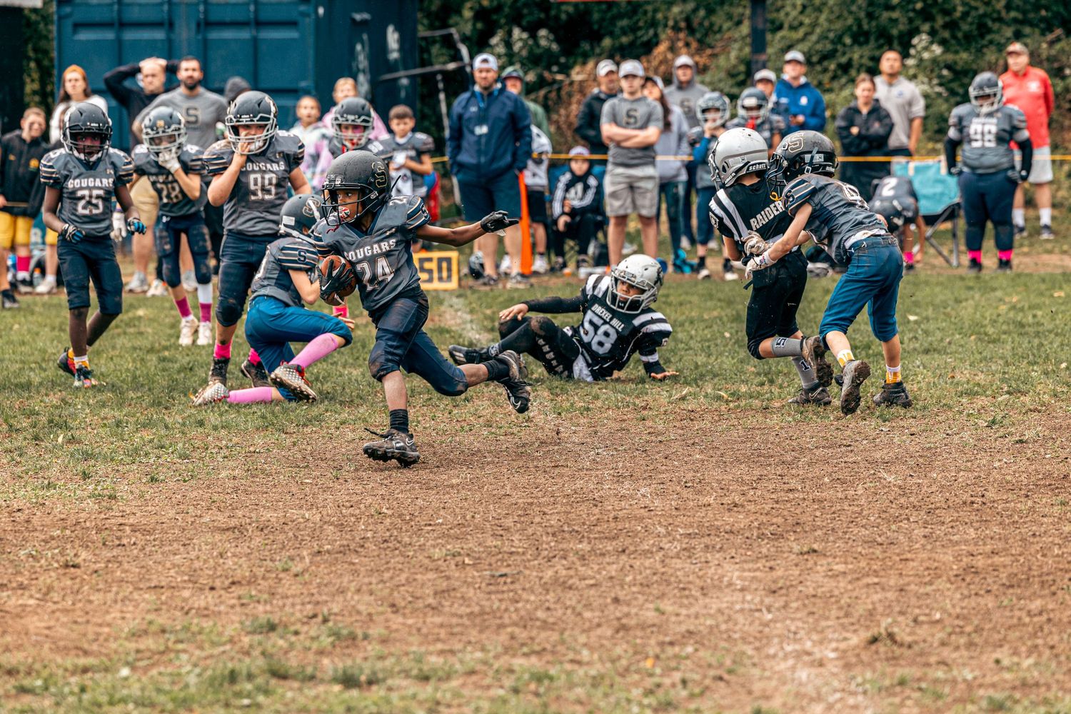 Youth football game action with players in dark uniforms making diving tackles on a dirt field.