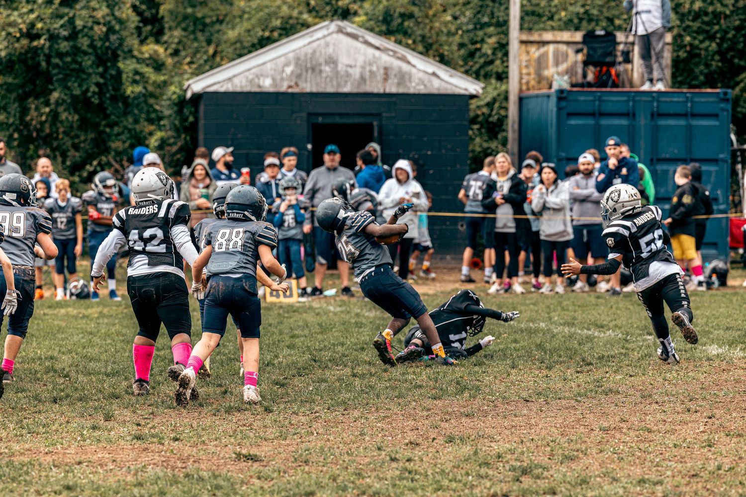 Youth football players in dark uniforms compete on a field with spectators and buildings in background.