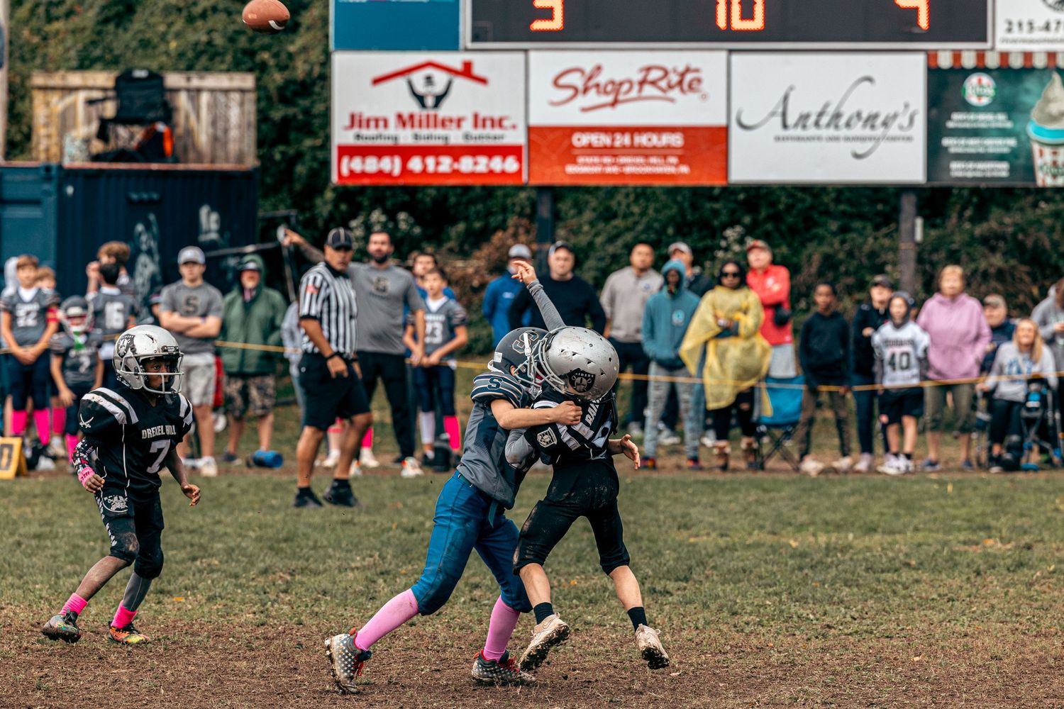 Youth football players engaged in a passing play with advertising banners visible in background.
