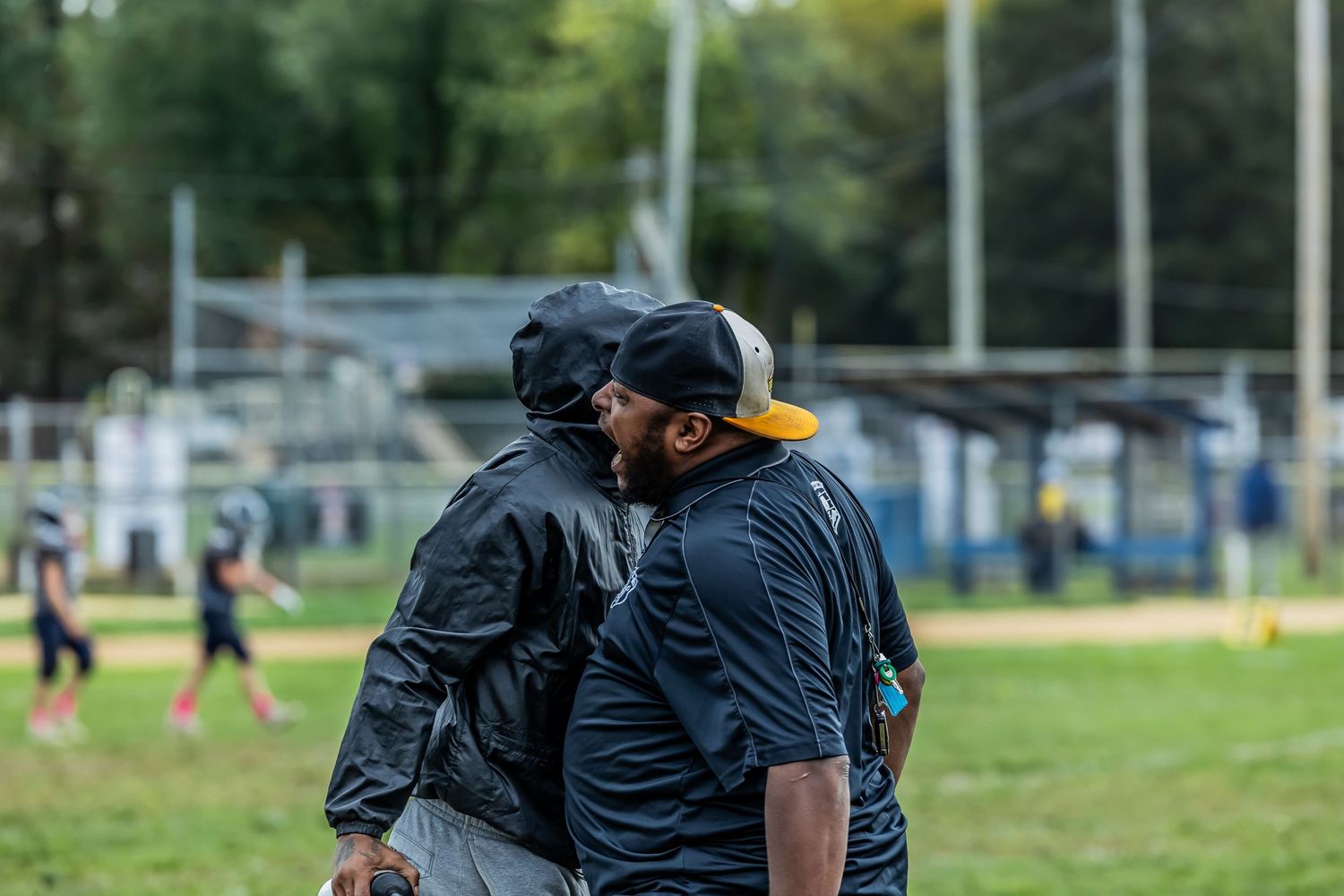 Two coaches in black jackets discuss strategy on the sidelines of a youth sports field.